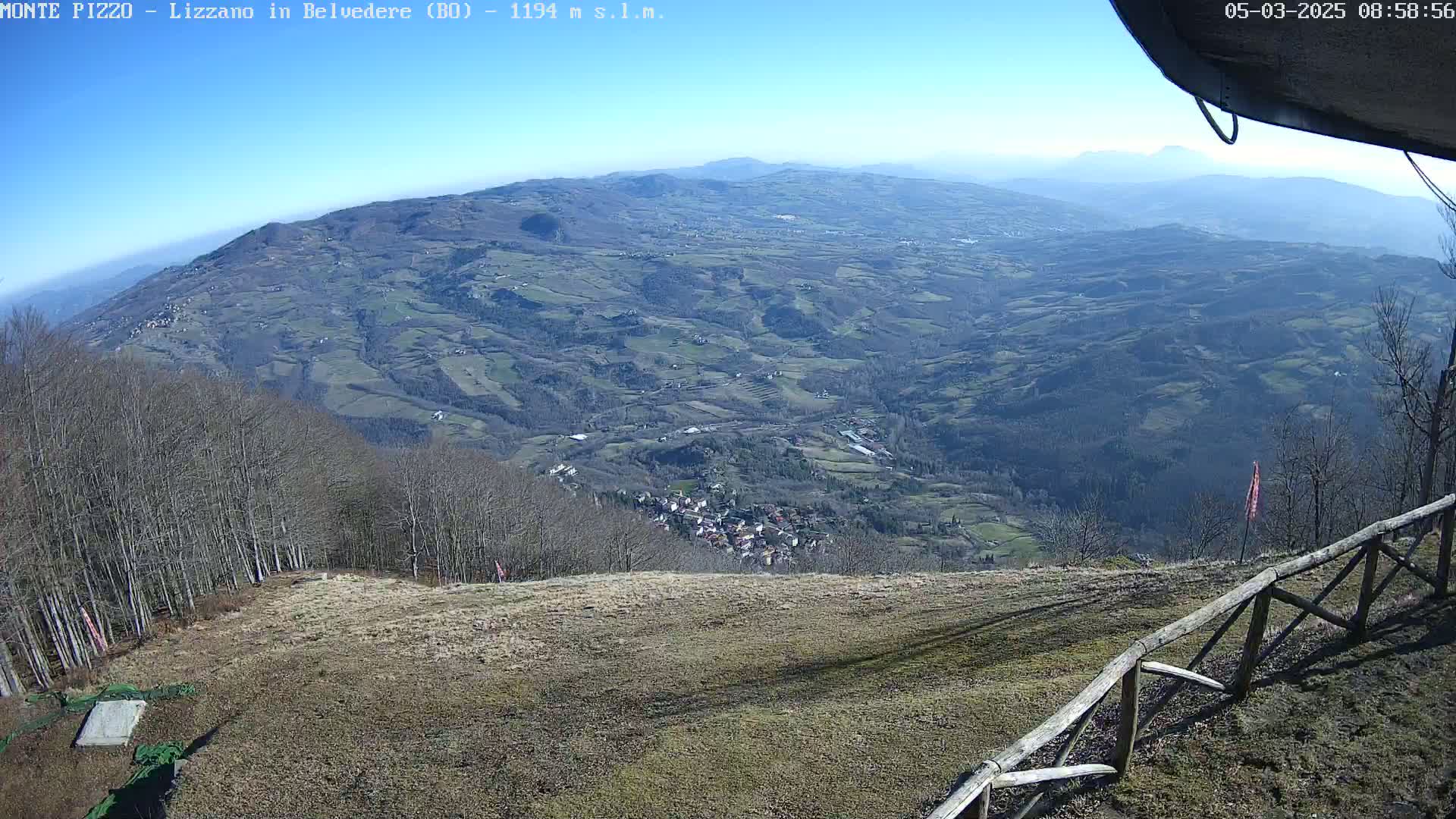 A panoramic view of a hilly landscape under a clear blue sky shows a village nestled in a valley, with bare trees and brown fields in the foreground.