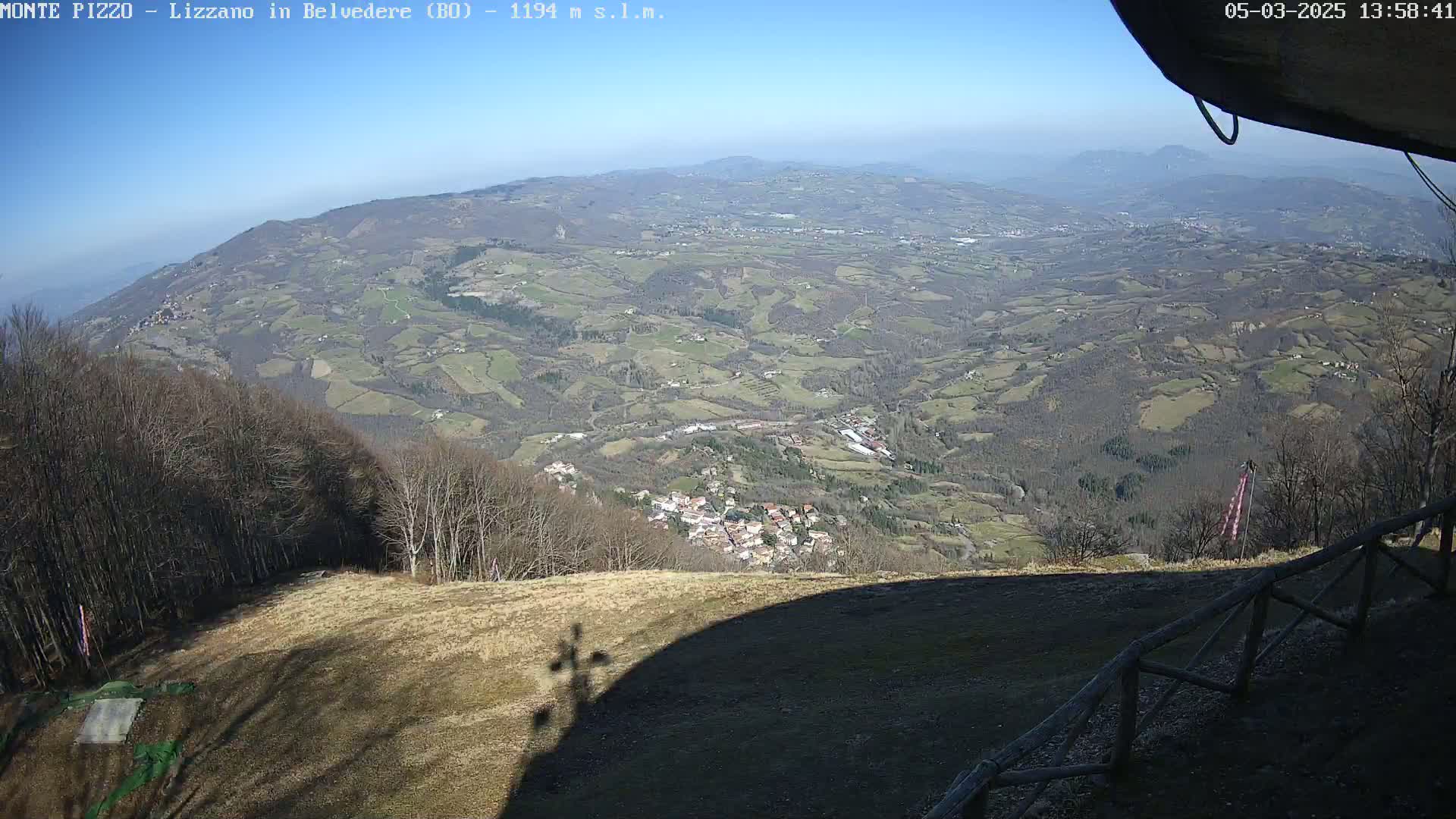 A high-angle, long shot on a sunny day shows a vast, hilly landscape dotted with small villages and farmland, viewed from a high vantage point with a wooden fence in the foreground.