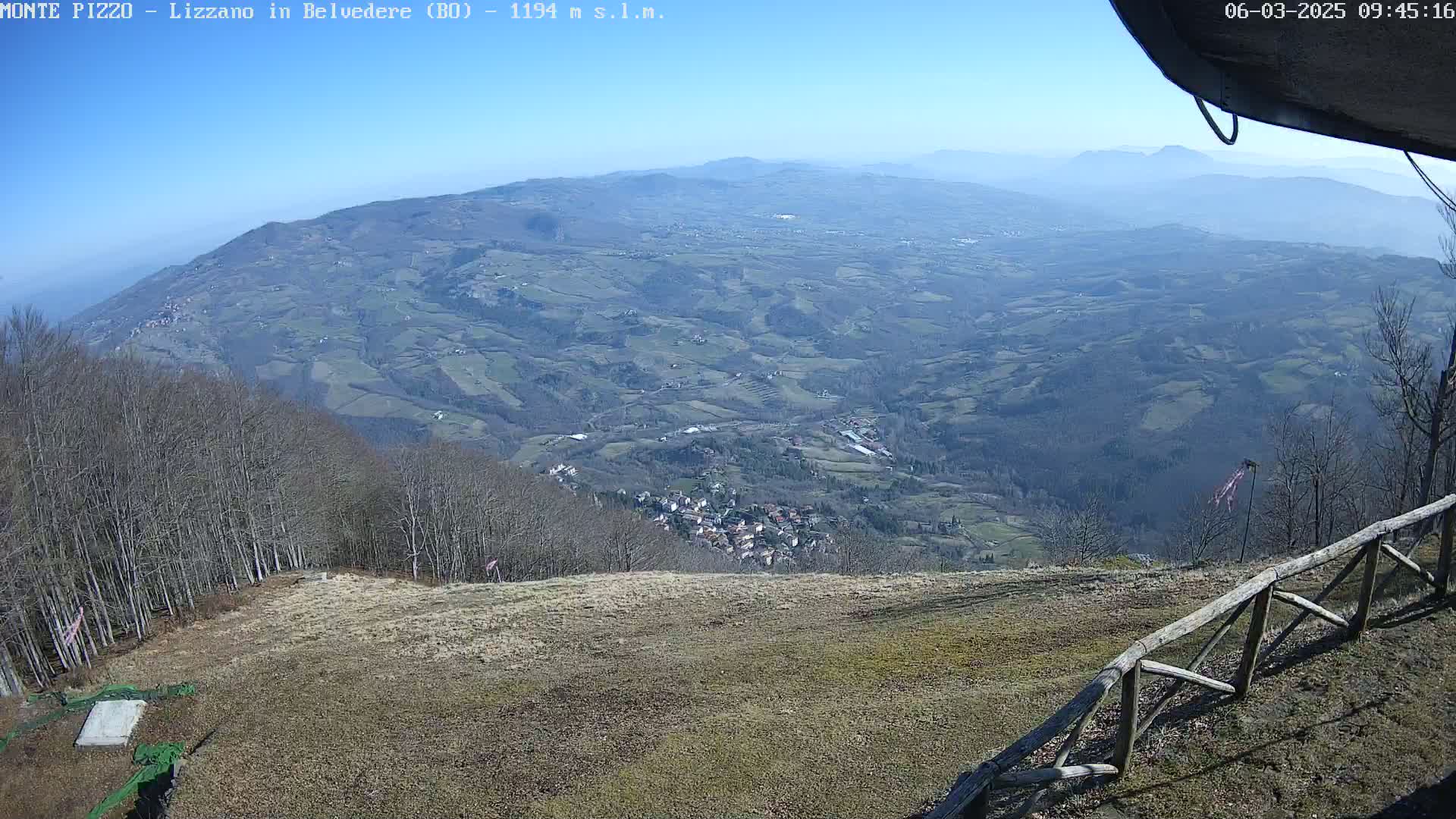 A high-angle, long shot on a clear, sunny day shows a vast, hilly landscape with a small village nestled among the trees, viewed from a high vantage point with a wooden fence in the foreground.