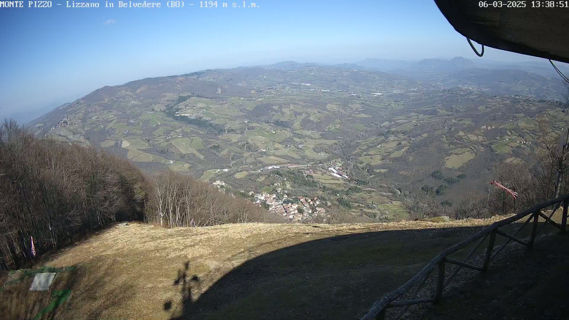 A high-angle, sunny view shows a vast, hilly landscape dotted with small towns and farmland, seen from a high vantage point with a wooden fence in the foreground.
