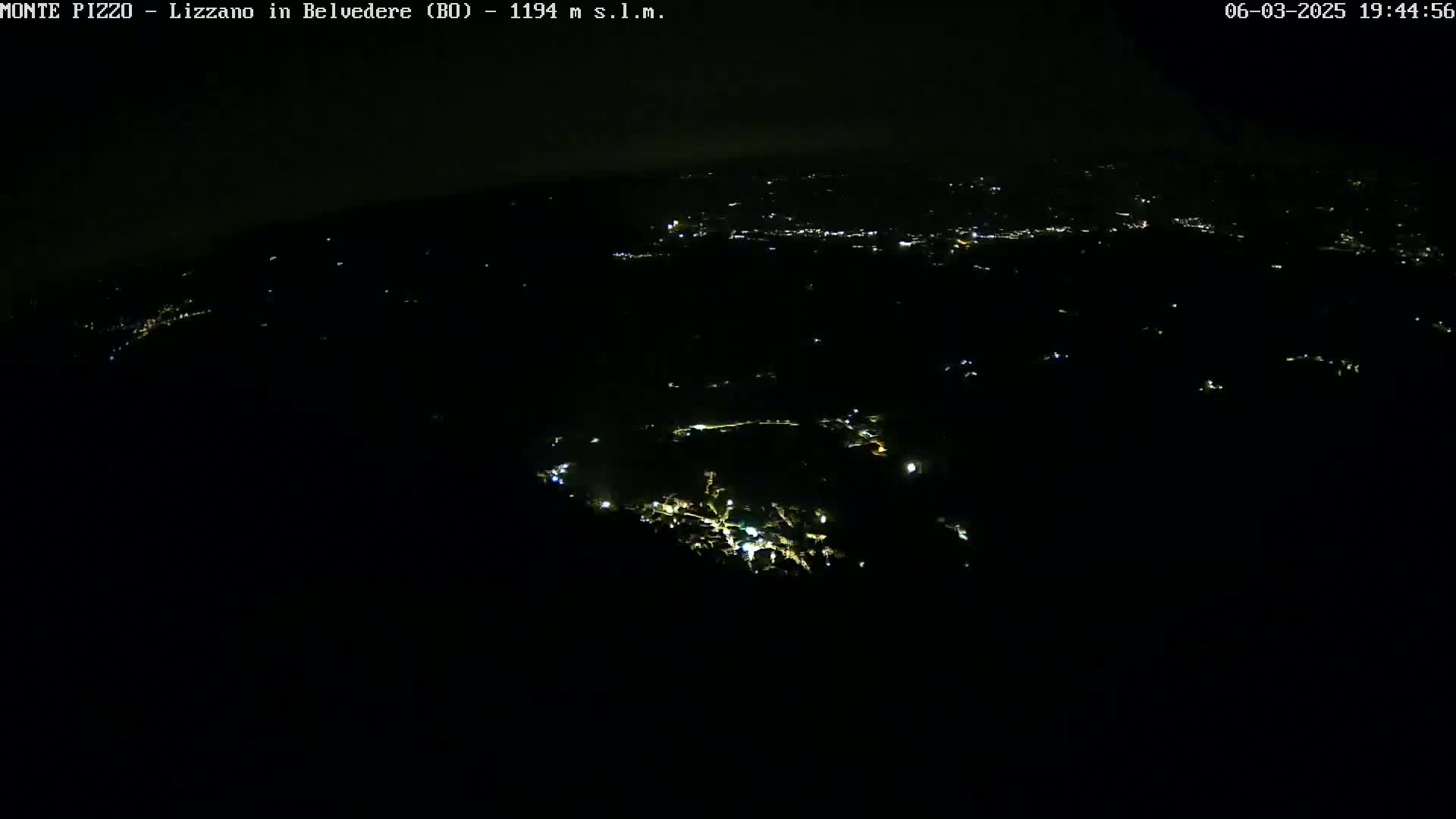 A nighttime, high-angle view shows a dark landscape speckled with scattered lights from a town or village below.