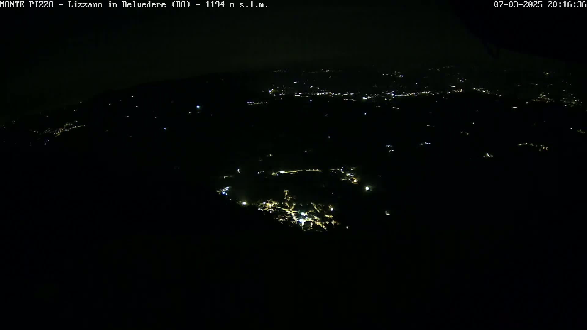 A nighttime, high-angle view shows a dark landscape sparsely dotted with the lights of a town or village.