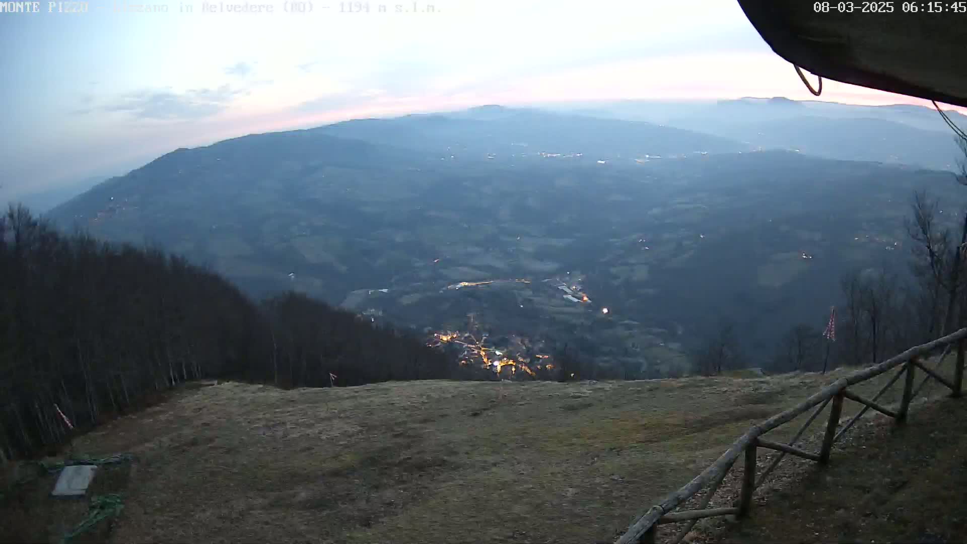A high-angle, twilight view shows a valley town nestled among hills and trees, under a hazy sky.