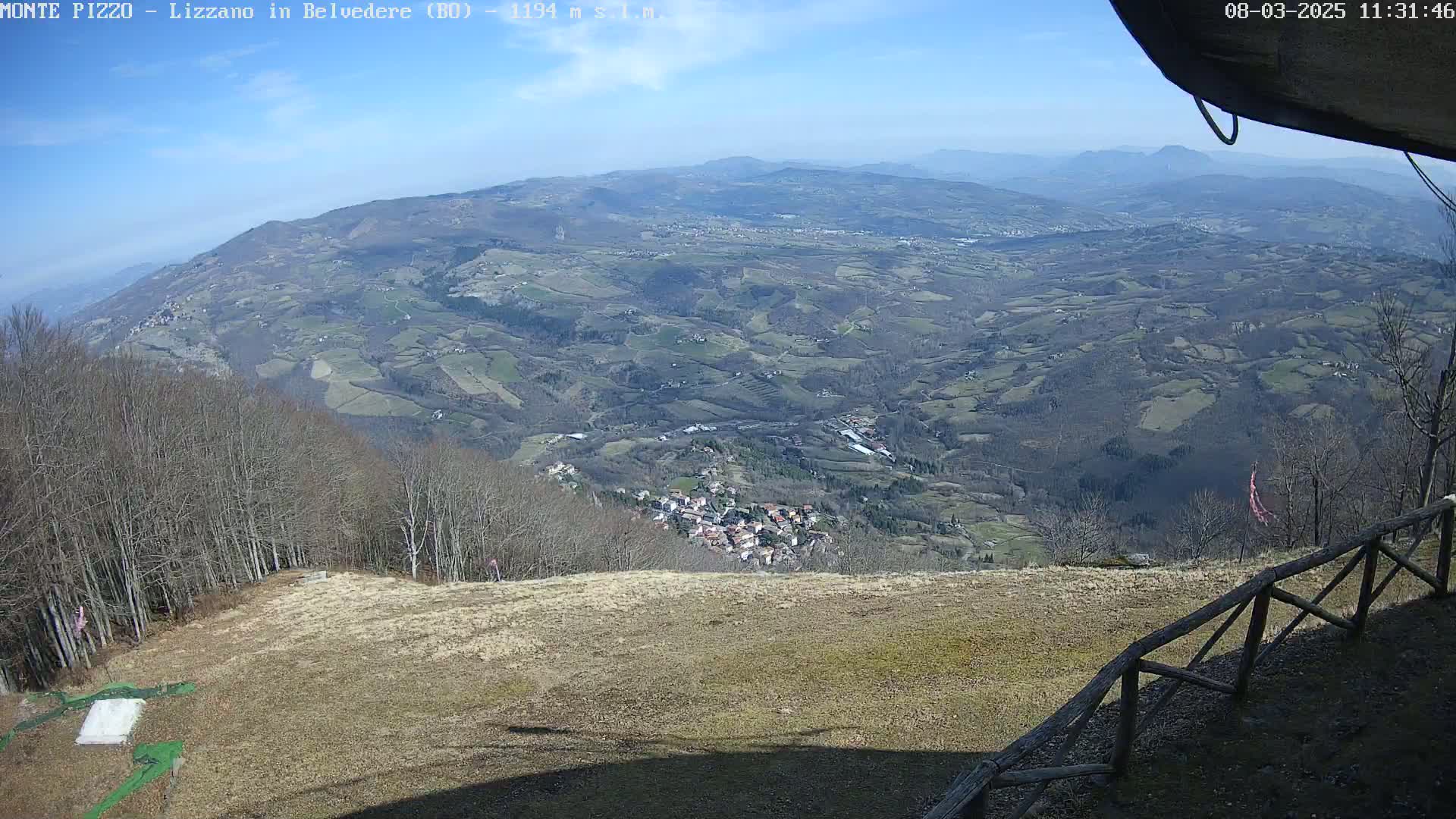 A high-angle view on a sunny day shows a vast, hilly landscape with a small town nestled in the valley below, surrounded by leafless trees and farmland.