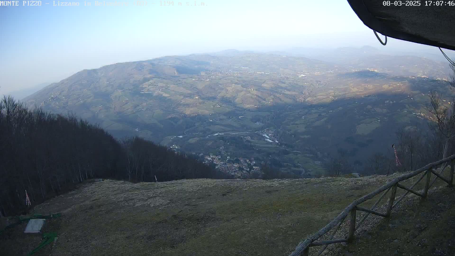 A high-angle, long shot shows a hazy, hilly landscape with a small town nestled in the valley below, viewed from a vantage point with a wooden fence in the foreground under a clear, light-blue sky.