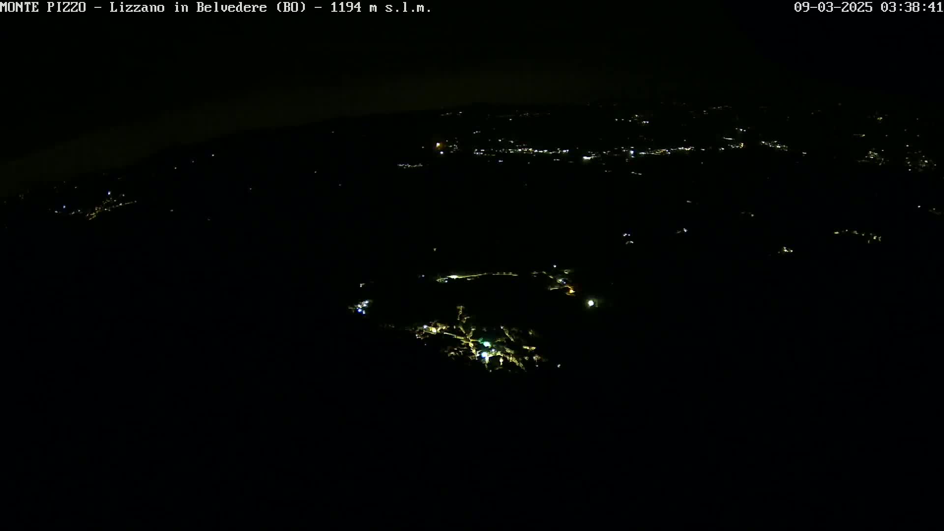 A nighttime, high-angle view shows a sparsely lit landscape with scattered lights from a town or village in the distance.