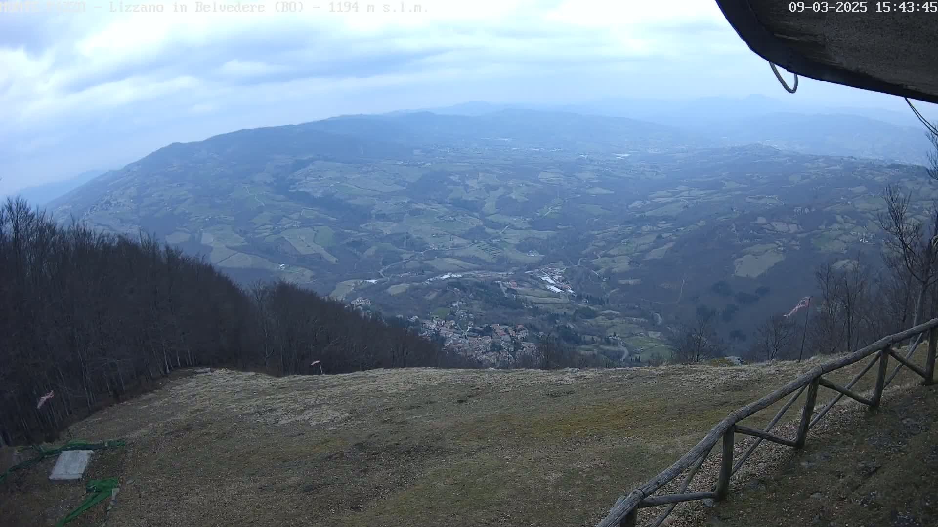 A high-angle, long shot of a sprawling, hilly landscape under a mostly cloudy sky, viewed from a high vantage point with a wooden fence in the foreground.