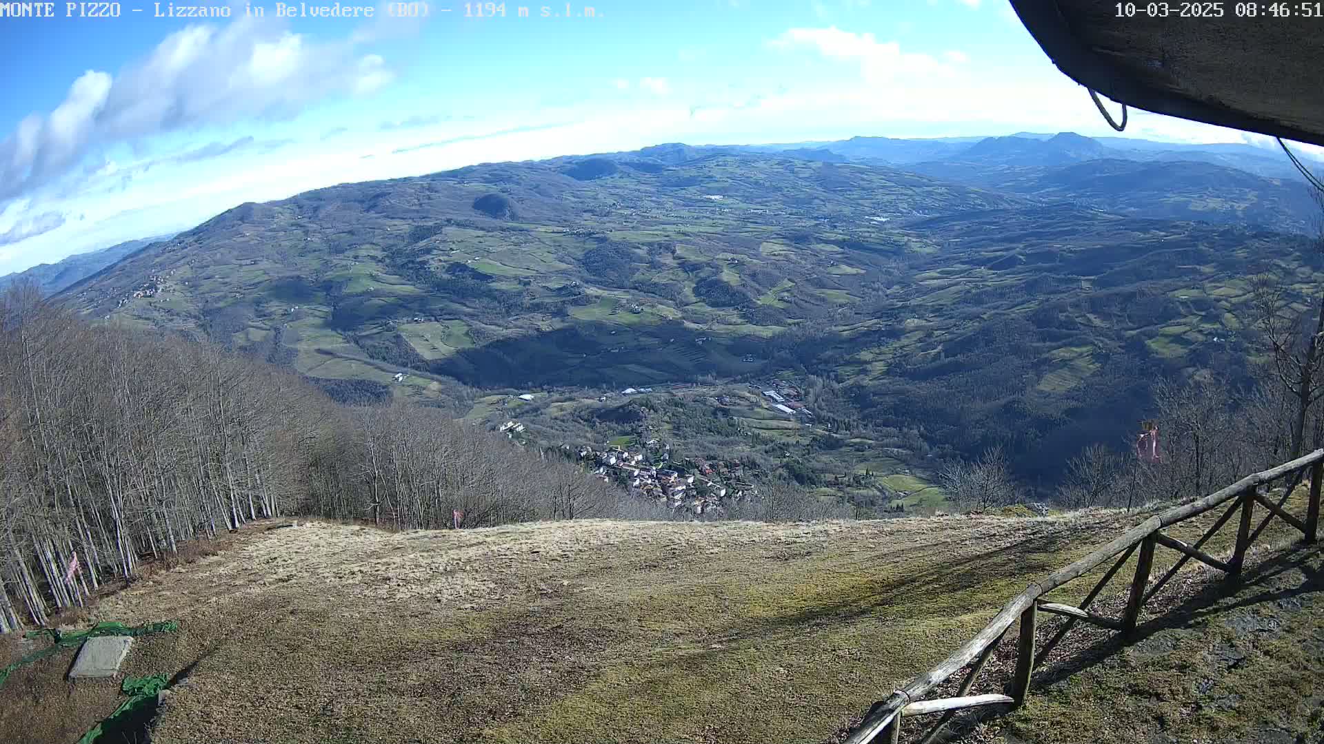 A high-angle view on a sunny day shows a sprawling valley with a small town nestled within rolling hills and a forest in the foreground.