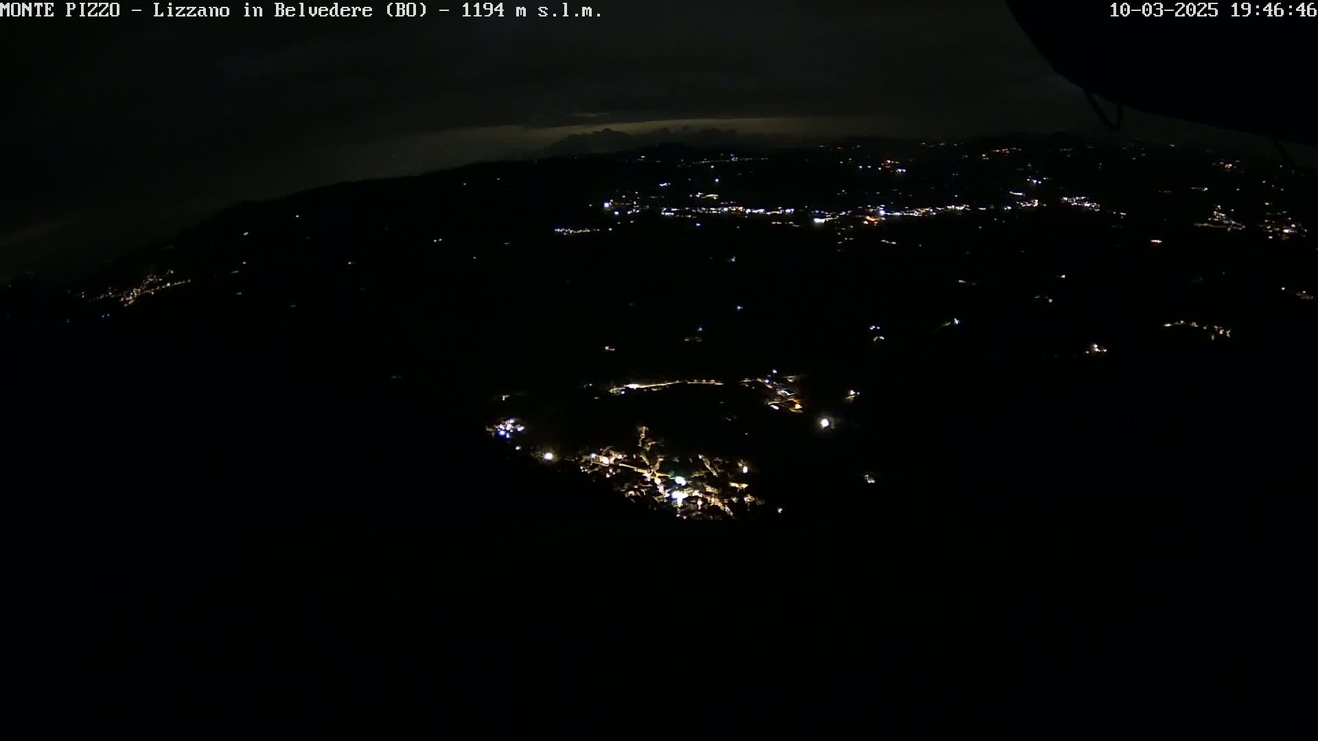 A nighttime, high-angle view shows a sprawling area with scattered lights, possibly a town or village, under a dark, cloudy sky.