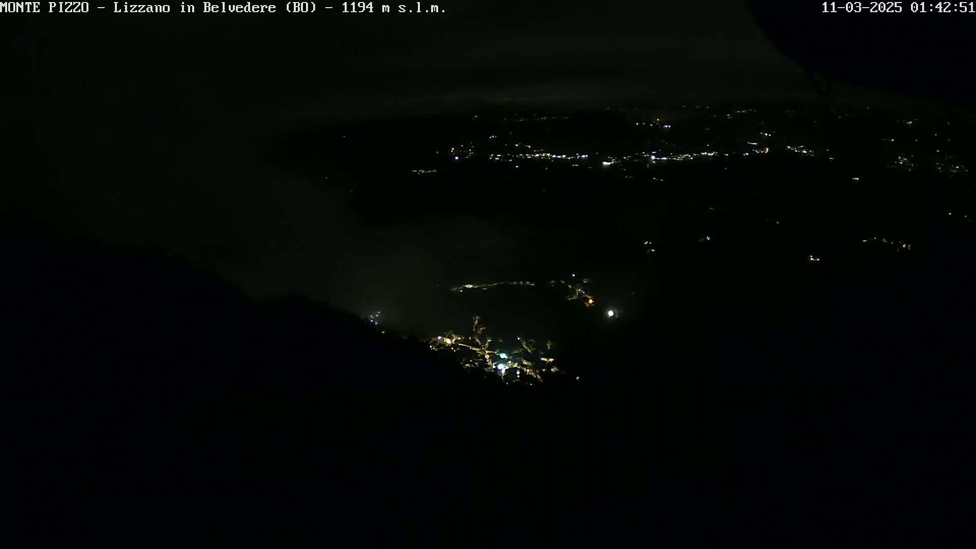 A nighttime, high-angle view shows a valley with scattered lights below, partially obscured by fog or low clouds.