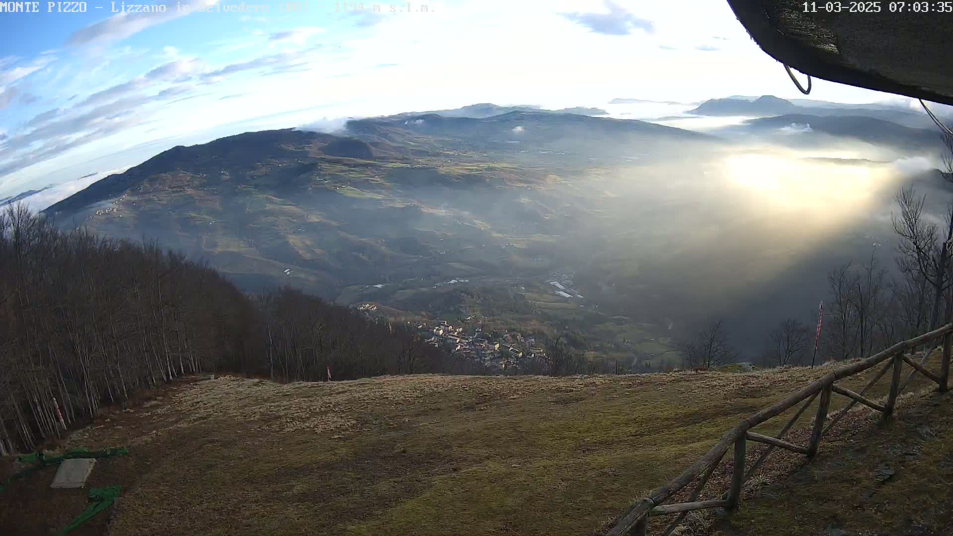 A hazy, sunlit valley stretches below a mountaintop vantage point, with a wooden fence and bare trees in the foreground.