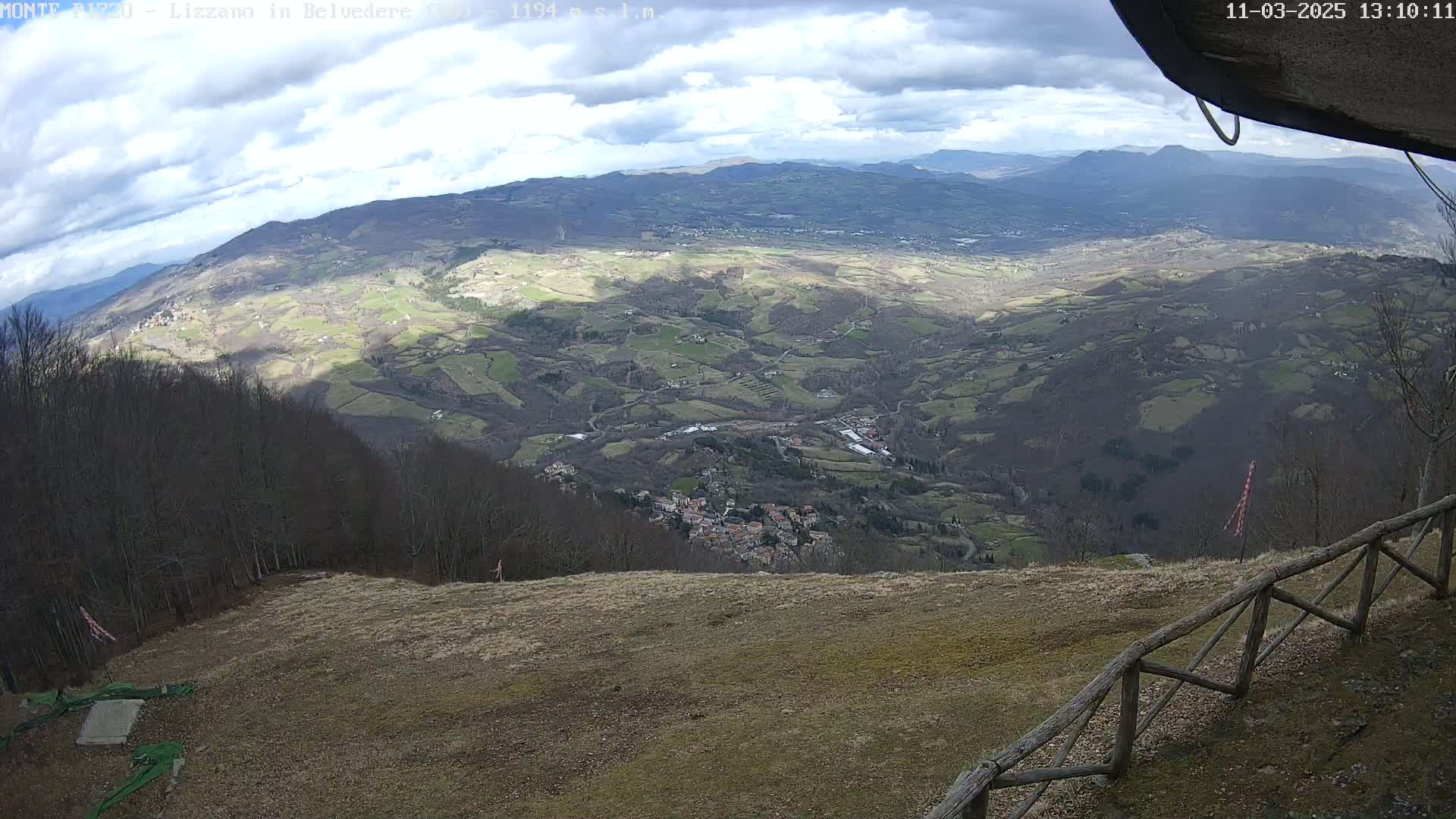 A high-angle view on a partly cloudy day shows a sprawling valley with a small town nestled among rolling hills and farmland, viewed from an elevated vantage point with a wooden fence in the foreground.