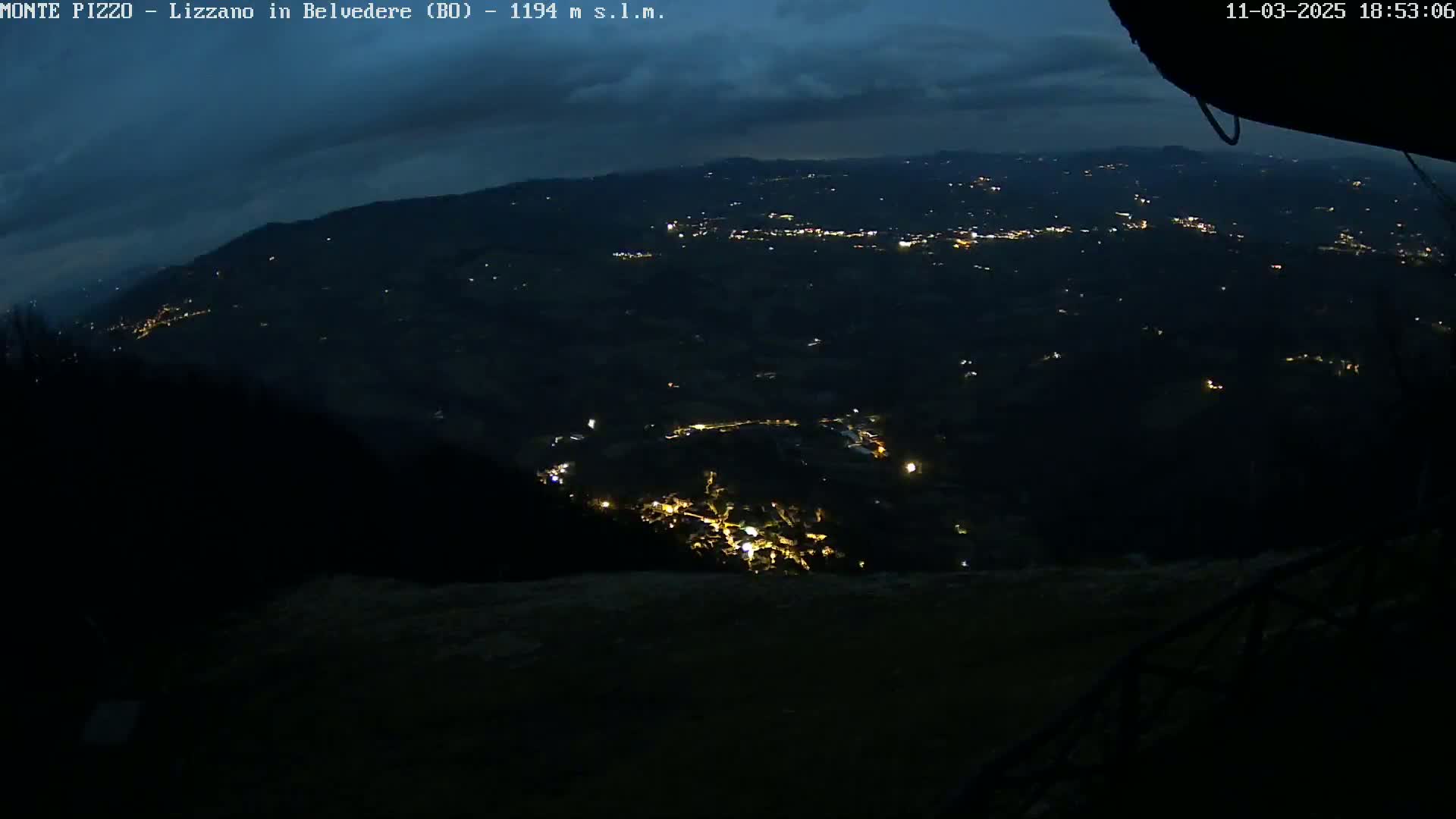 A nighttime, high-angle view of a valley with scattered lights, under a dark, cloudy sky.