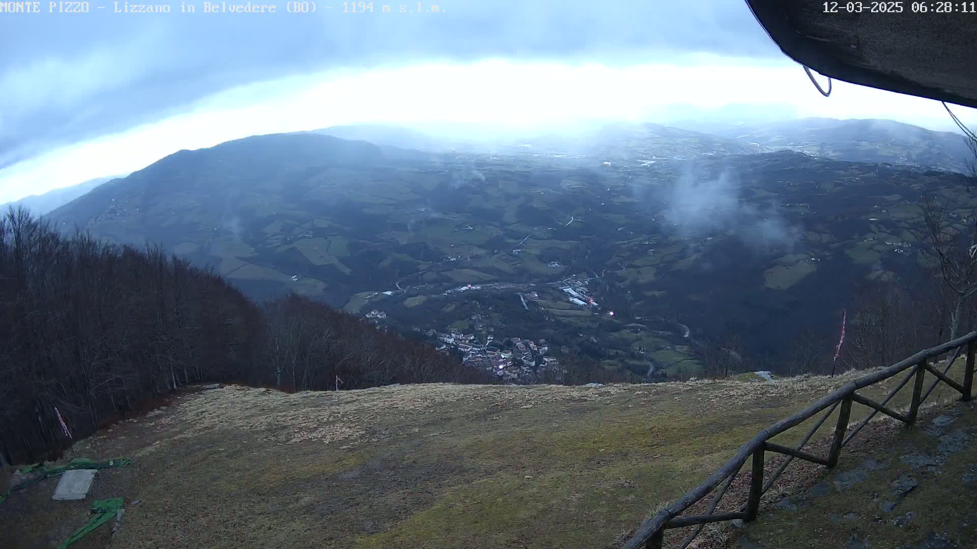 A high-angle, long shot shows a valley town nestled amongst rolling hills and mountains on an overcast day, viewed from a high vantage point with a wooden fence in the foreground.