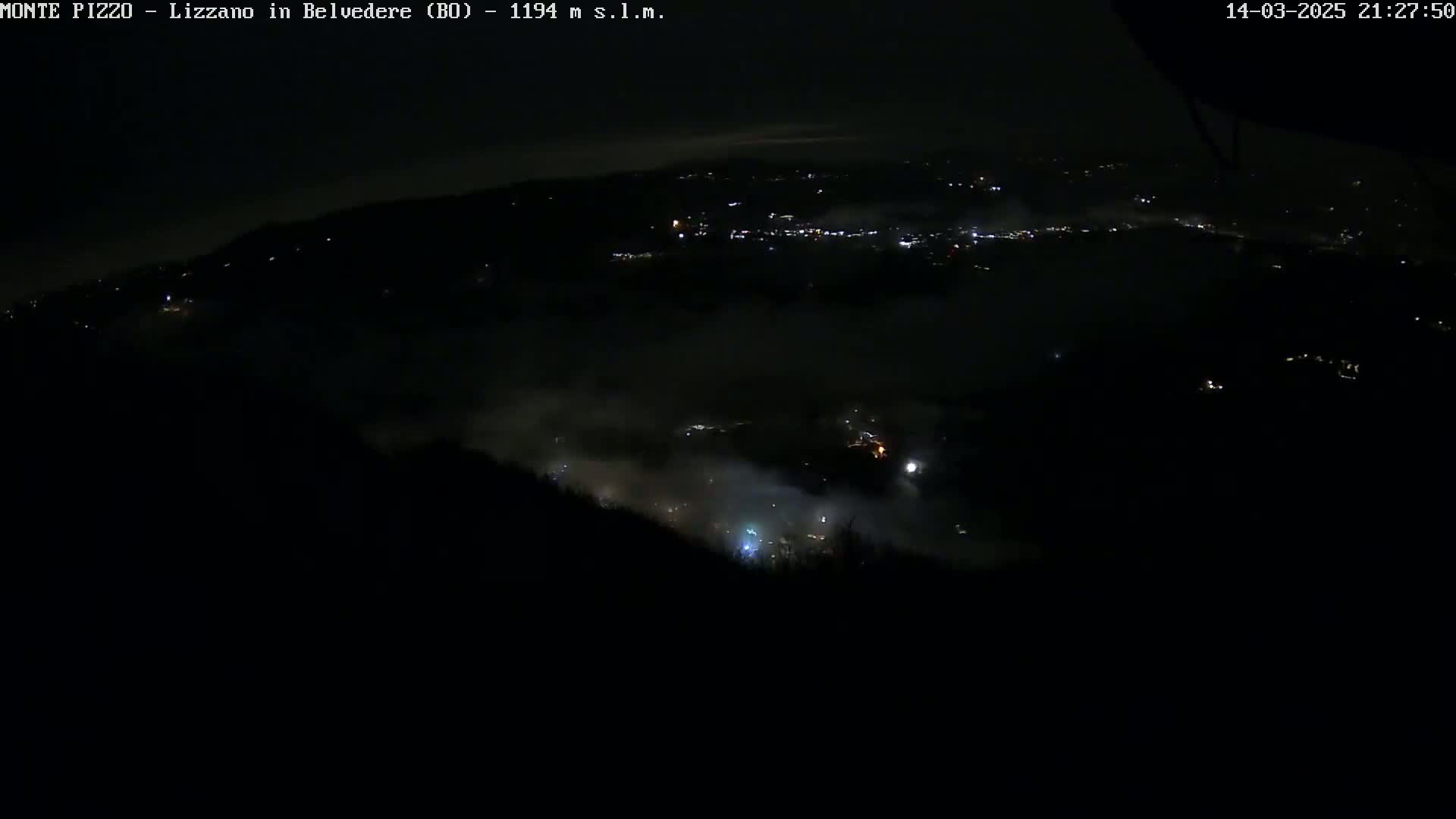 A nighttime, high-angle view shows a valley shrouded in fog, with scattered lights visible in the distance.