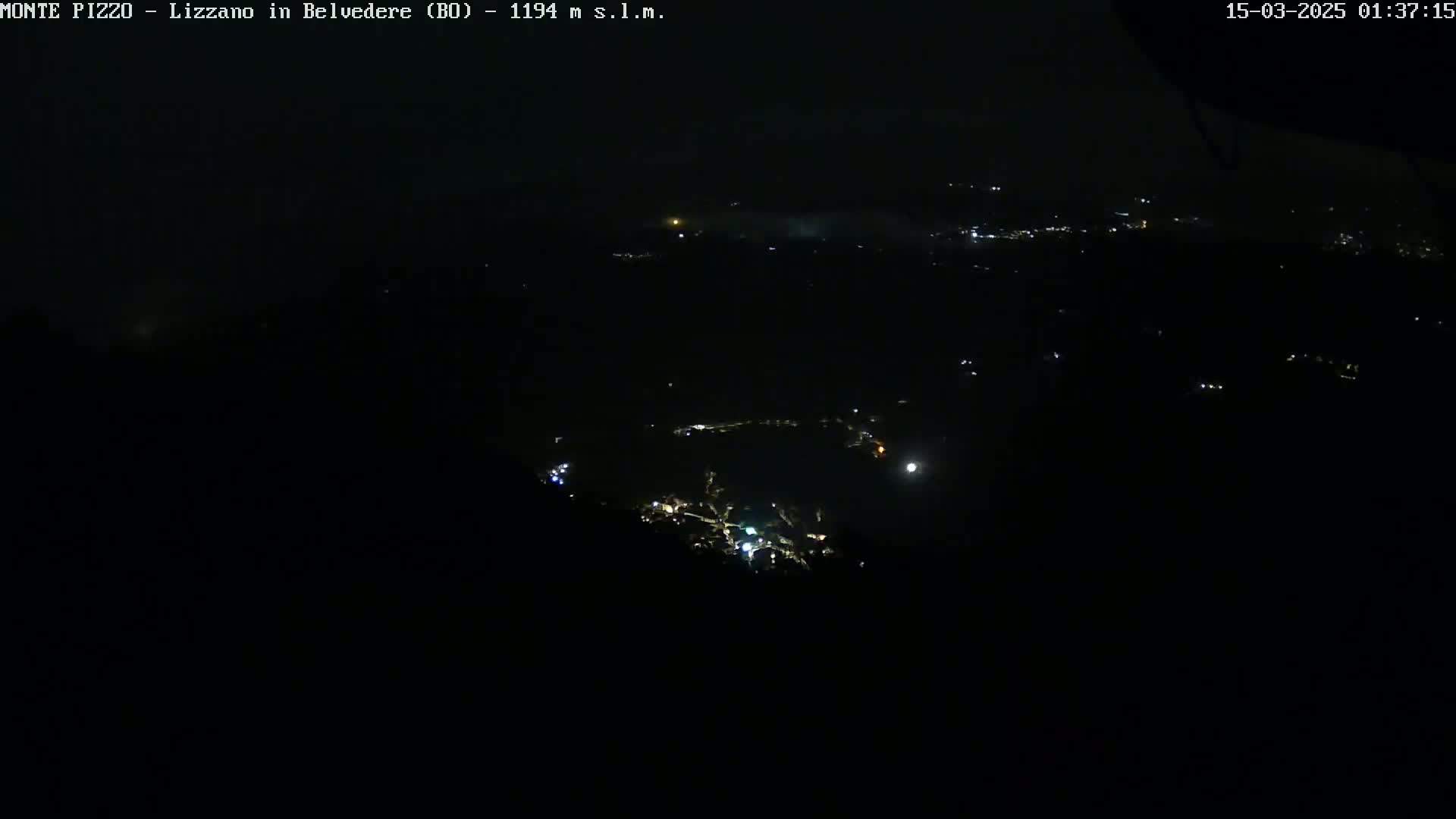 A nighttime, high-angle view shows a dark landscape with sparsely scattered lights, possibly a town or village, partially obscured by fog or low clouds.