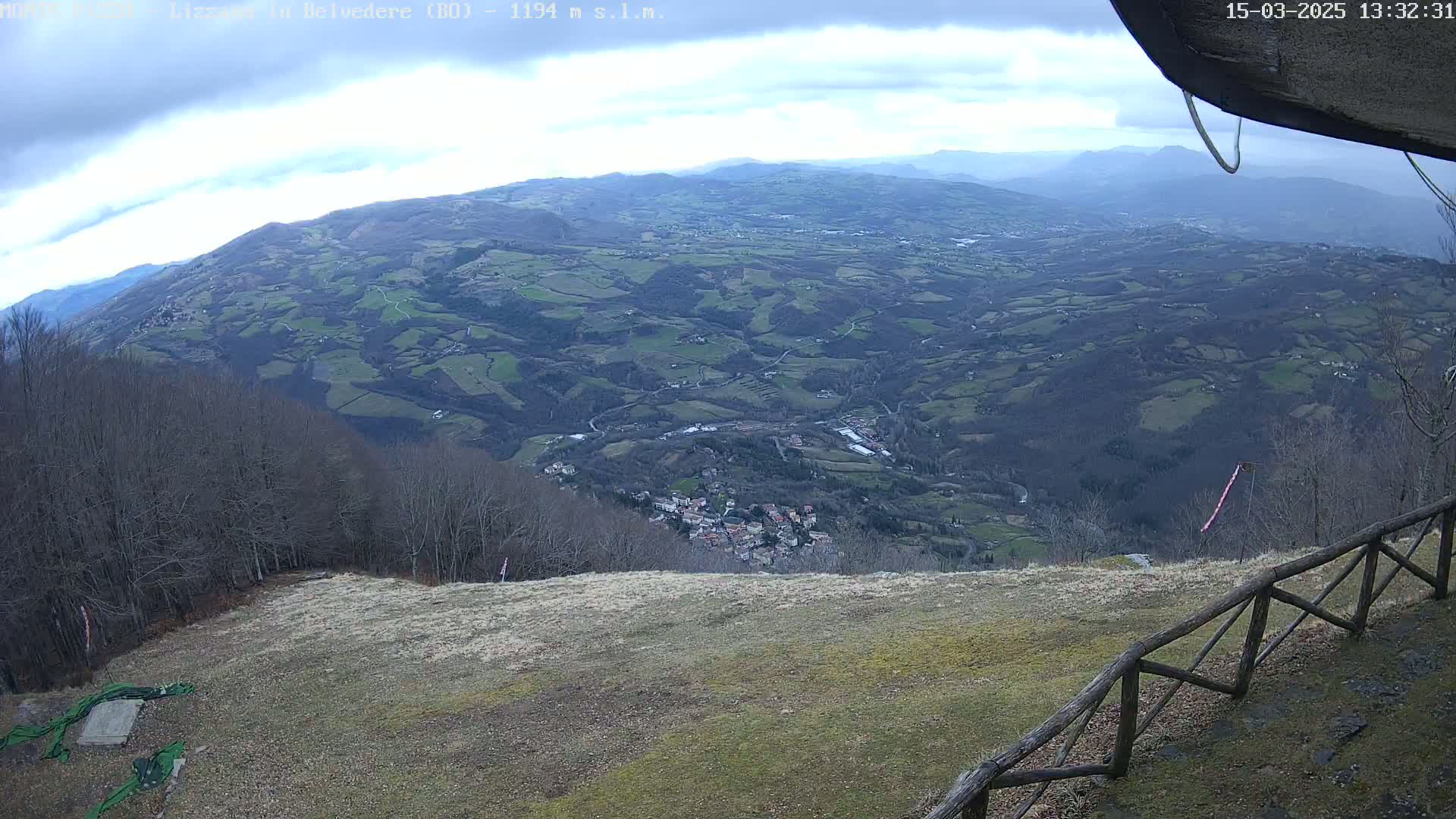 A high-angle view on an overcast day shows a sprawling valley with a small town nestled amongst rolling hills and forests, observed from a high vantage point with a wooden fence in the foreground.