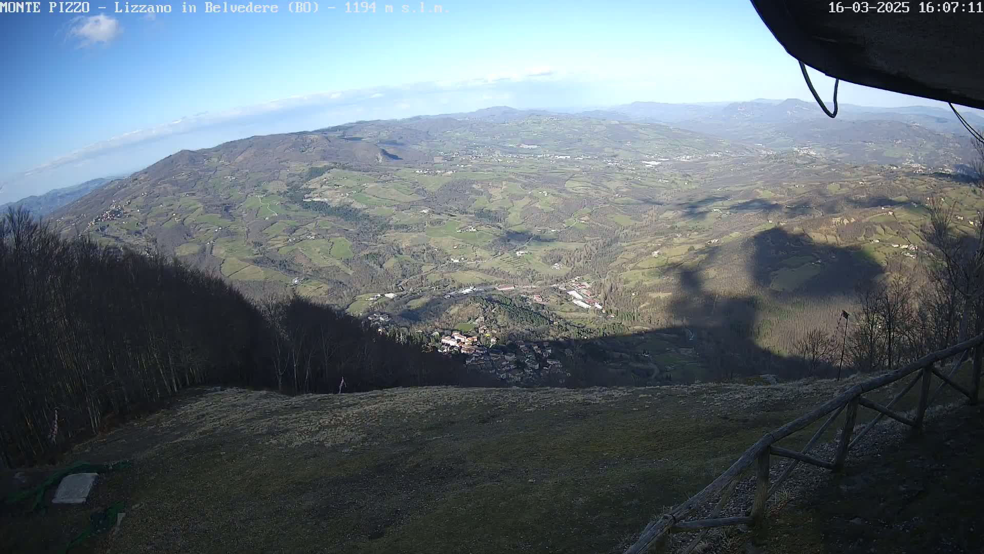 A vast, hilly landscape stretches out under a clear blue sky, with a small village nestled in the valley below, partially obscured by a large shadow cast from a nearby structure.