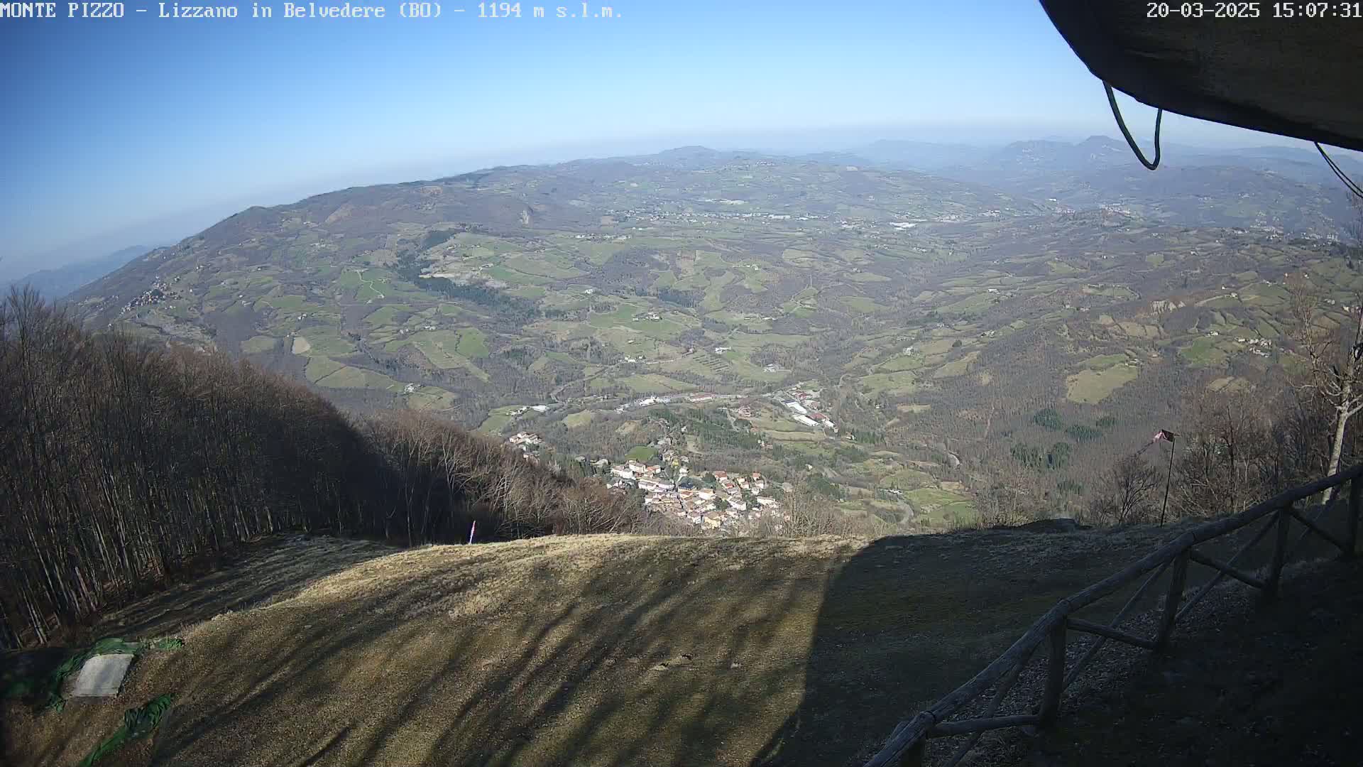 A high-angle view on a clear day shows a vast expanse of rolling hills and a small village nestled within a valley, observed from a wooded mountaintop with a wooden fence in the foreground.