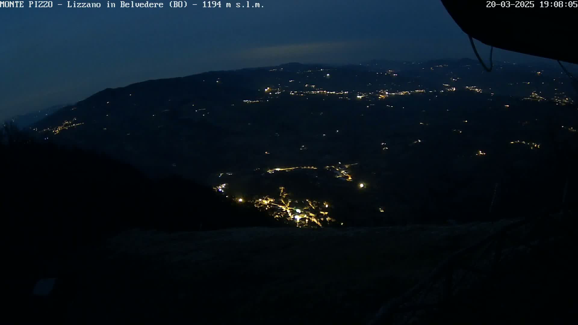 A nighttime, high-angle view shows a sprawling landscape of dimly lit towns and villages nestled among dark hills under a dusky sky.
