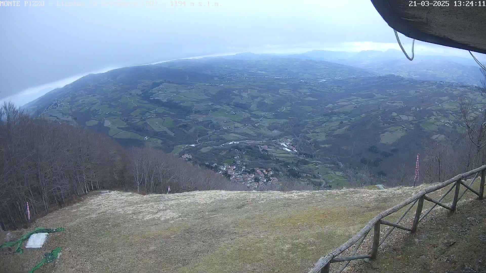 A high-angle view shows a vast, hilly landscape under an overcast sky, with a wooden fence and sparse, dry grass in the foreground.