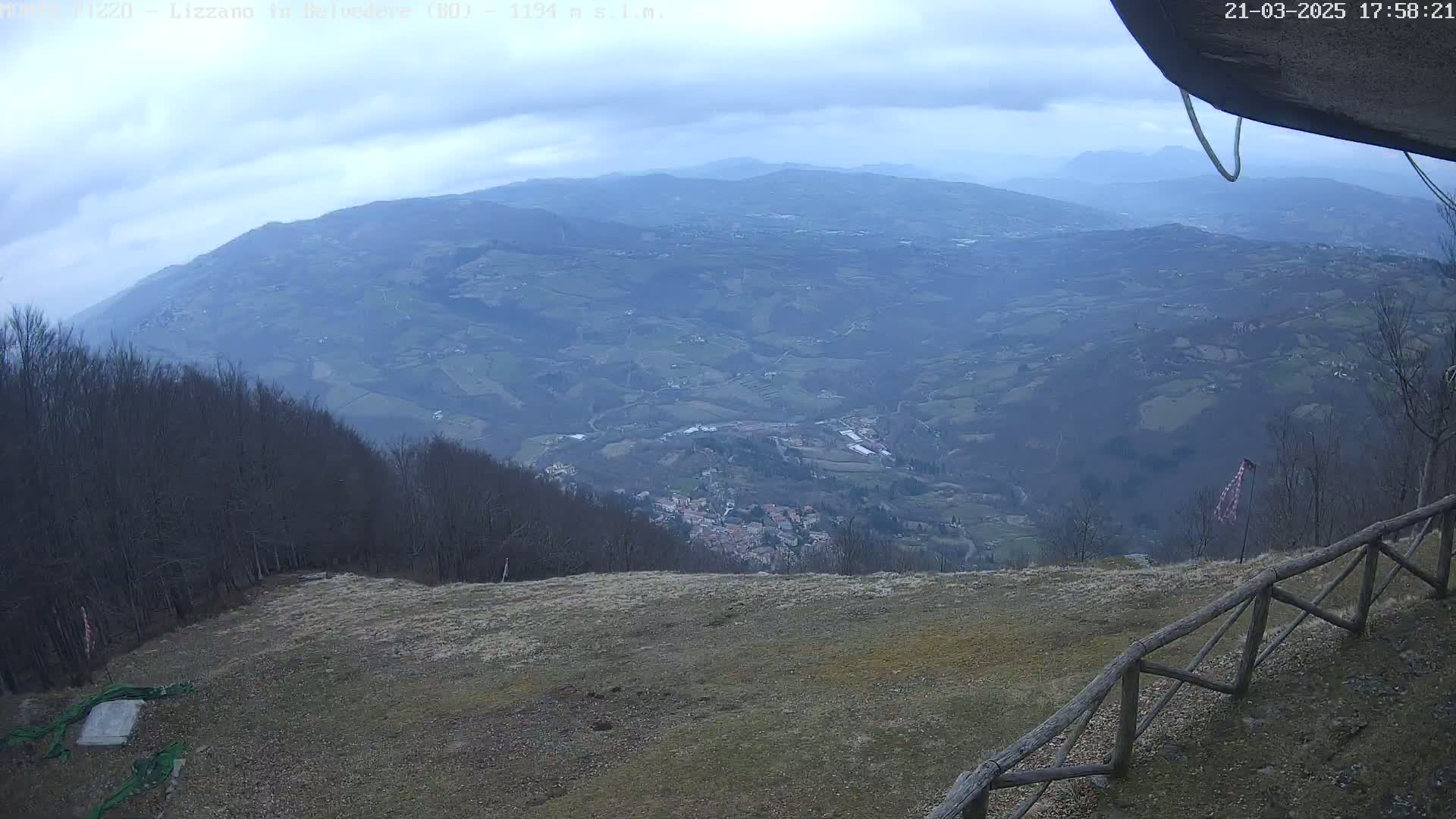 A high-angle view on an overcast day shows a sprawling valley with rolling hills and a small town nestled within, all overlooked from a vantage point with a wooden fence.