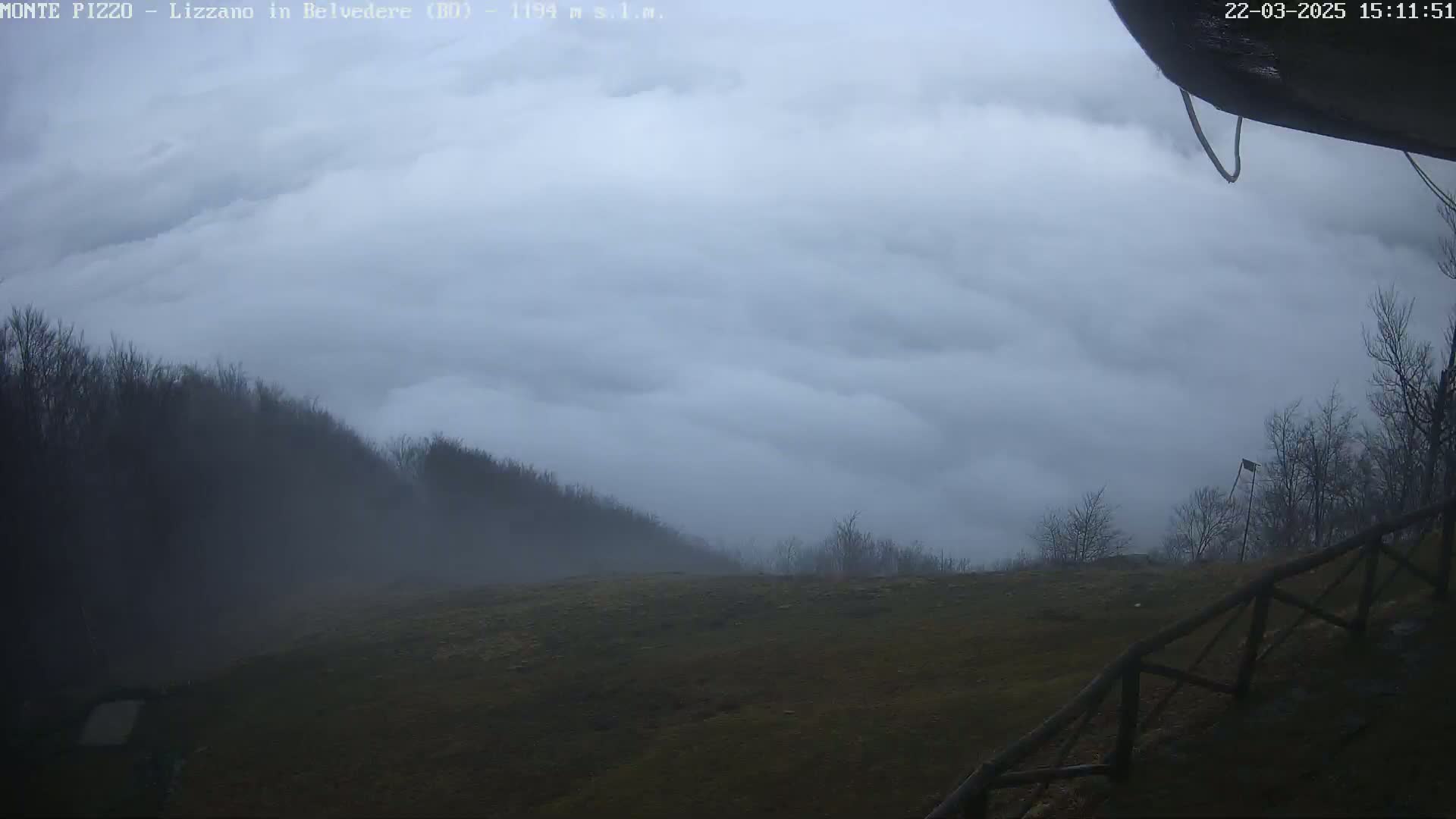 A foggy landscape shows low-lying clouds covering hills and trees, with a grassy field in the foreground and a wooden fence in the lower right corner.