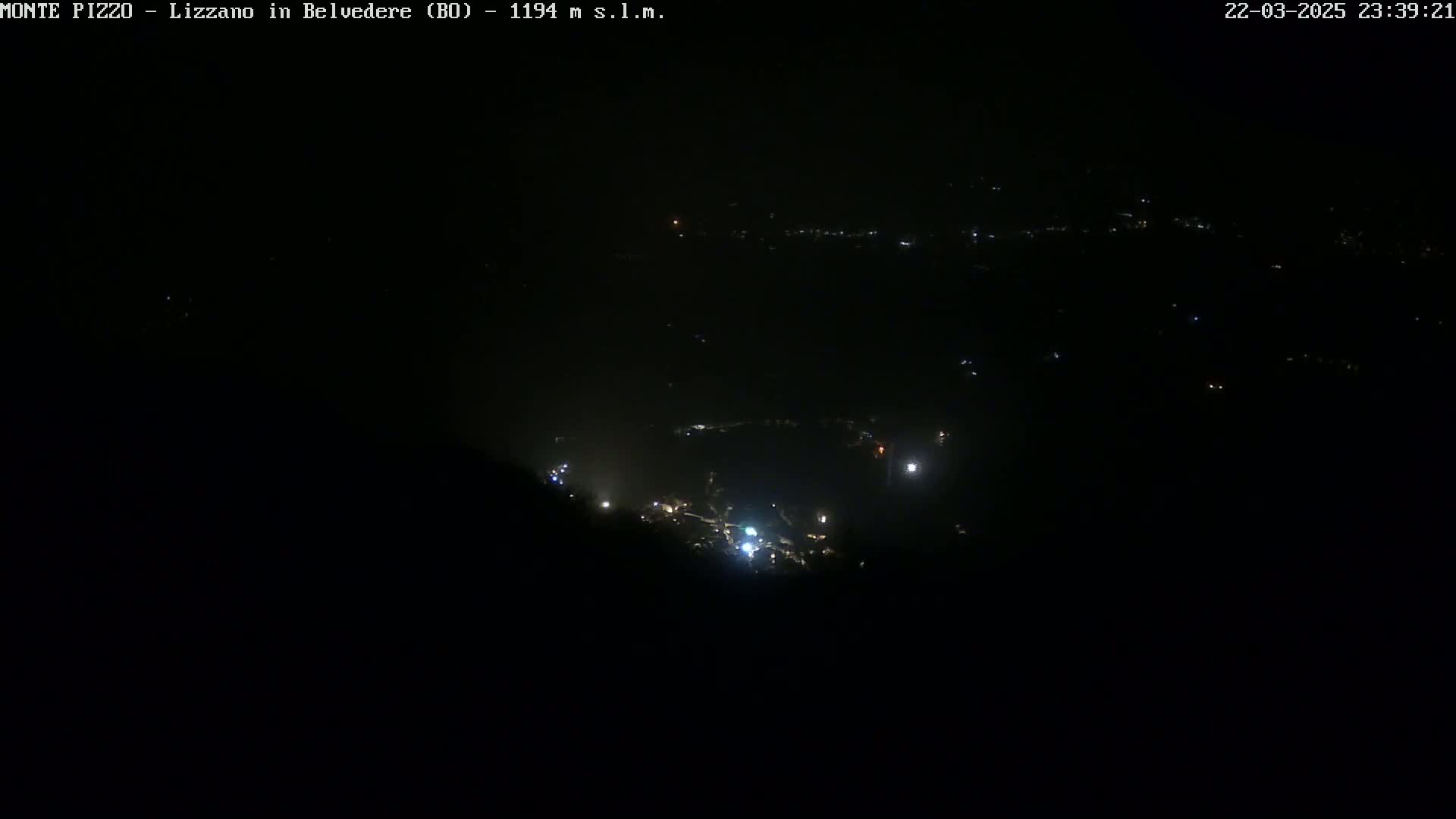 A nighttime, high-angle view shows a dark landscape sparsely dotted with faint lights from a distant town or village.