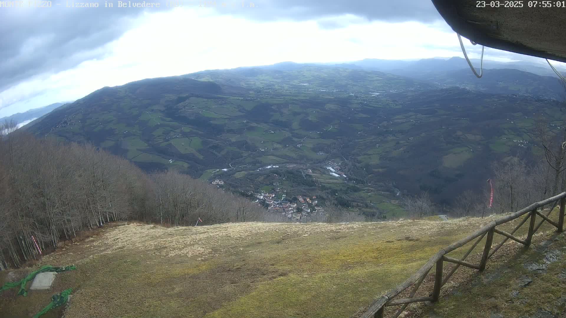 A high-angle view shows a sprawling valley of farmland and a small village under a cloudy sky, seen from a grassy hillside with a wooden fence in the foreground.