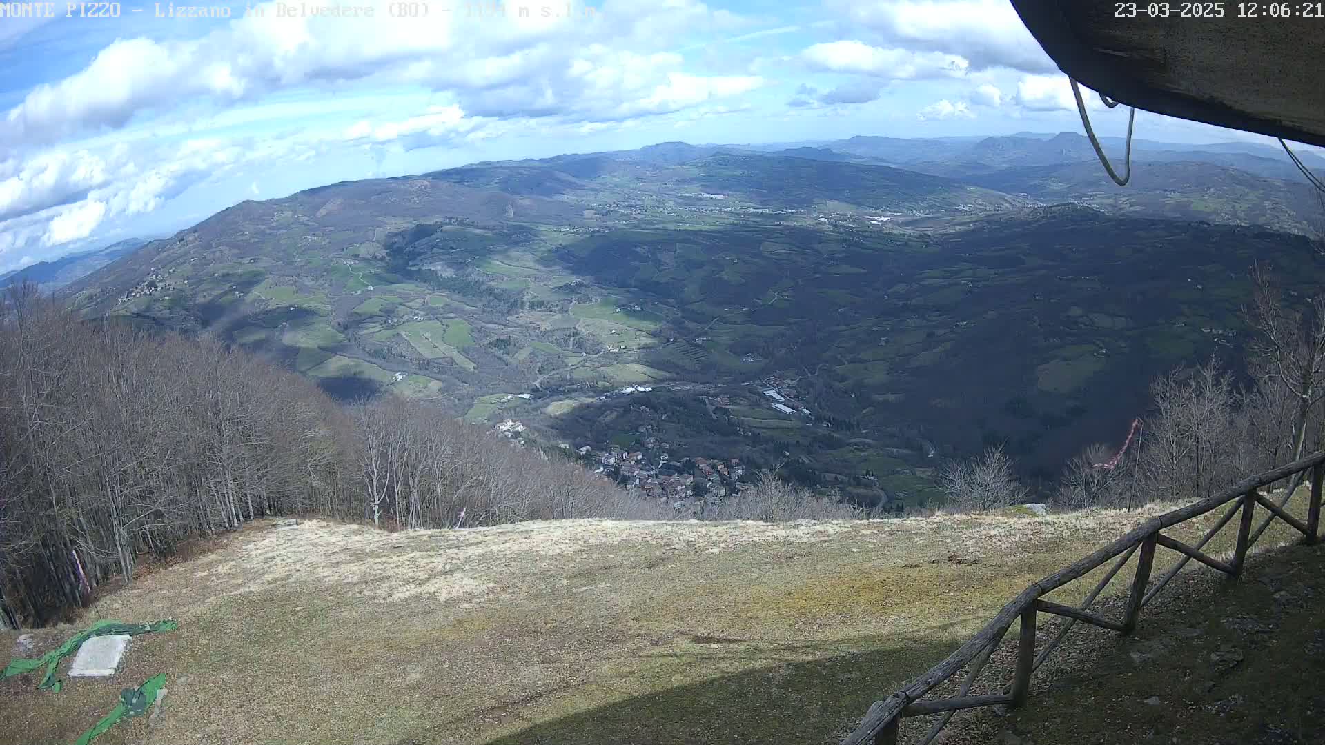 A high-angle view on a partly cloudy day shows a sprawling valley with a small town nestled amongst rolling hills and forests, seen from a vantage point overlooking a grassy area with a wooden fence.
