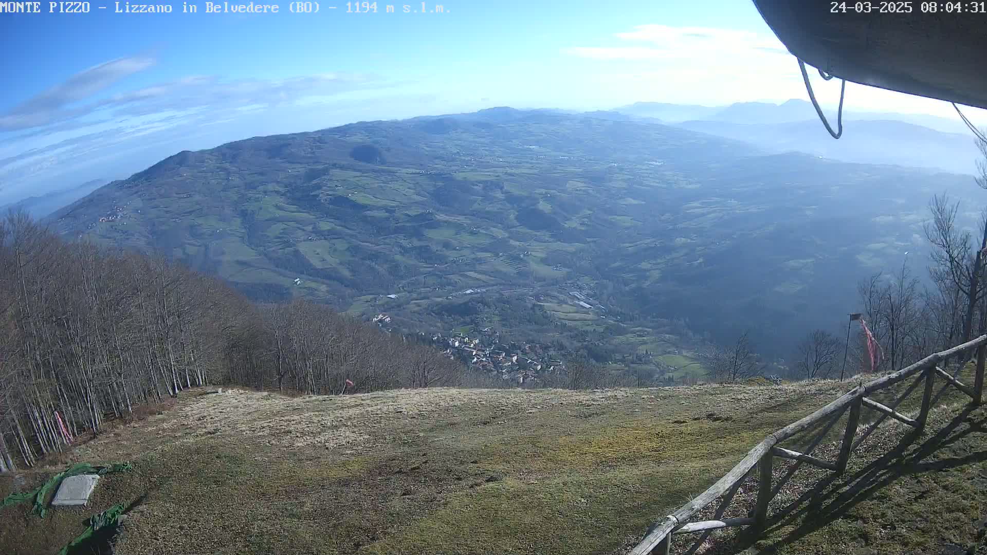 A high-angle view on a clear, sunny day shows a vast mountainous landscape with a town nestled in a valley below, all viewed from a grassy area with a wooden fence in the foreground.