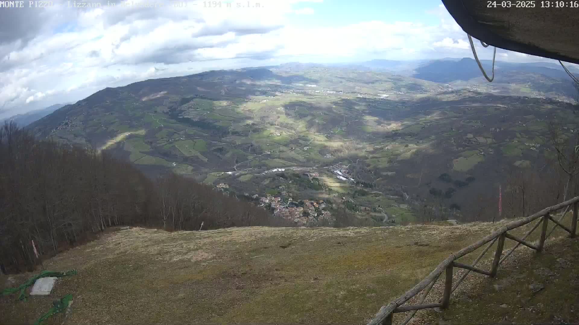 A high-angle view on a partly cloudy day shows a vast expanse of rolling hills and valleys, with a small village nestled below, all seen from a high vantage point with a wooden fence in the foreground.