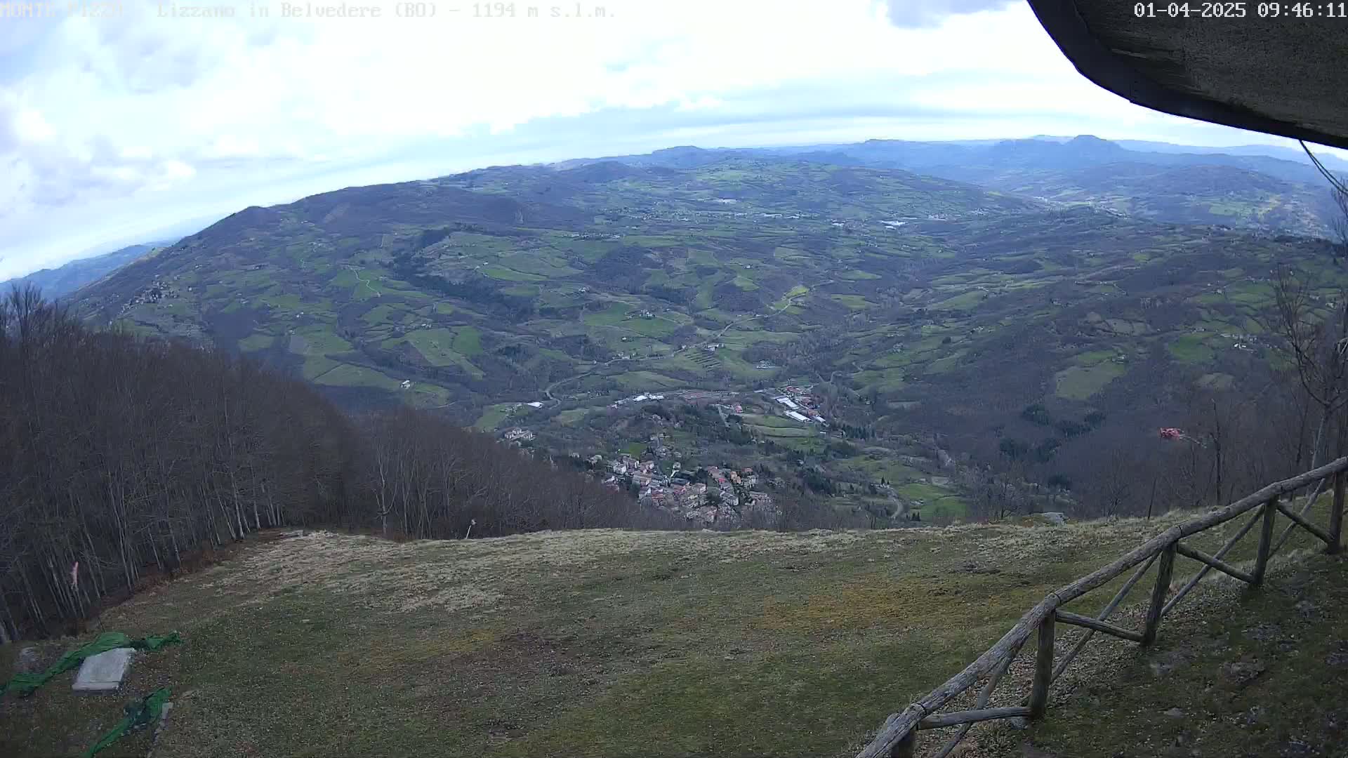A high-angle view shows a vast, hilly landscape under a partly cloudy sky, with a small village nestled in the valley below and a wooden fence in the foreground.