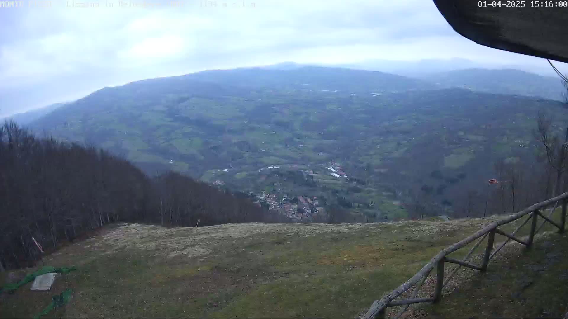 A high-angle view on an overcast day shows a mountainous valley with a small village nestled within, viewed from a grassy hilltop with a wooden fence.