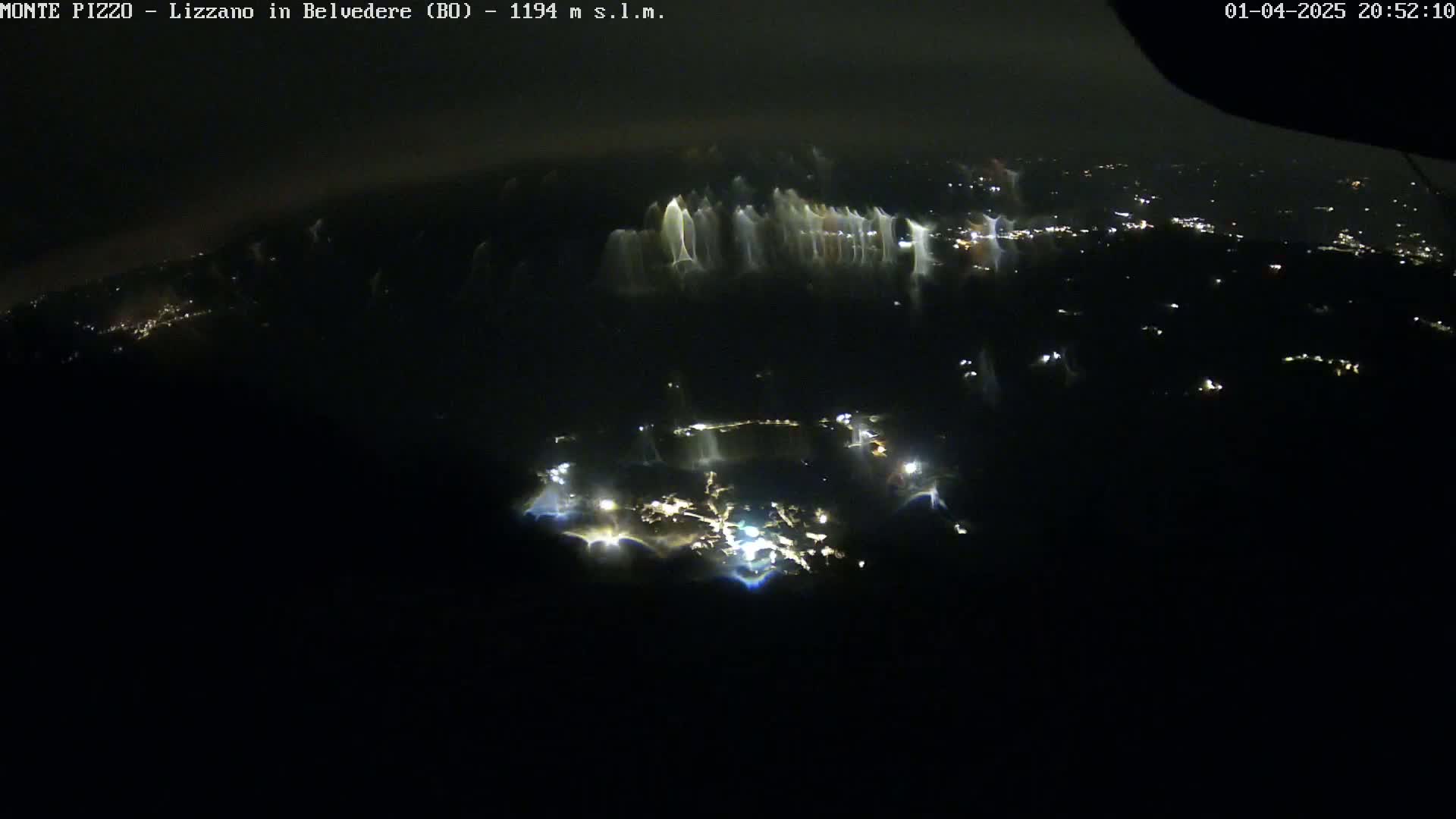 A nighttime aerial view shows a sprawling town illuminated by scattered lights, with a hazy or cloudy atmosphere.
