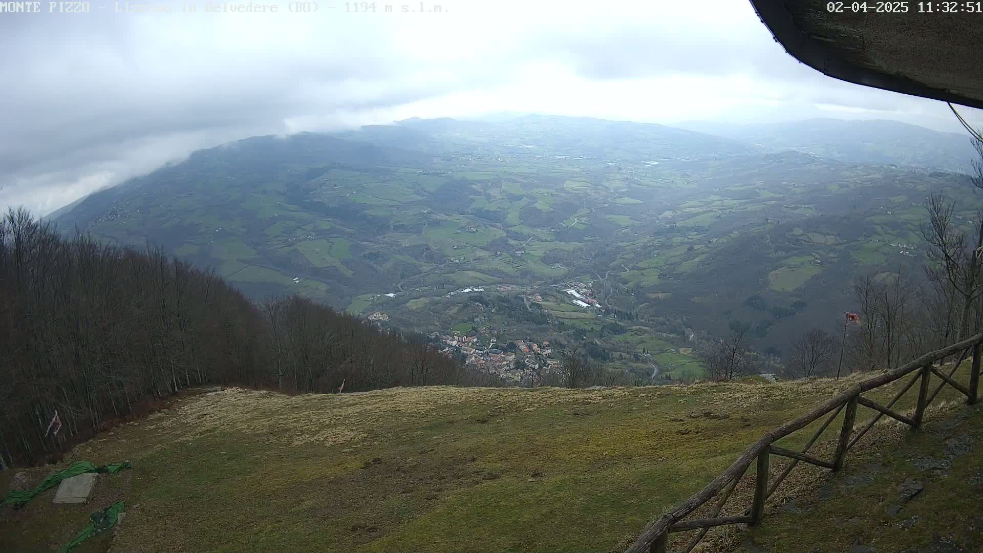 A high-angle view on an overcast day shows a sprawling valley with a small town nestled among rolling hills and farmland, viewed from a grassy hilltop with a wooden fence.