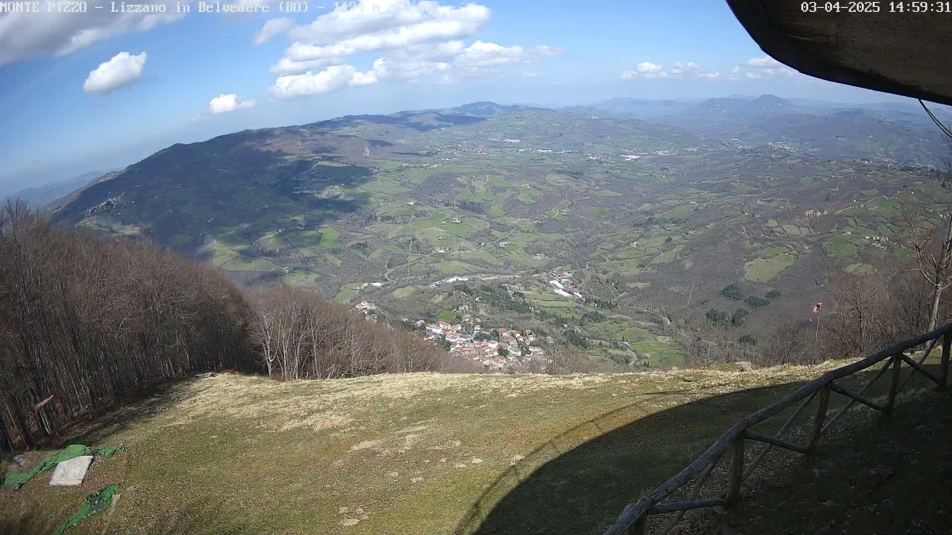 A high-angle view of a partly sunny valley dotted with small towns and farmland, seen from a mountaintop with a wooden fence in the foreground.