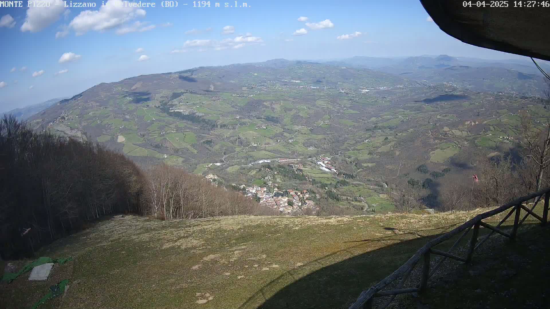 A high-angle, sunny view shows a sprawling valley with a small village nestled among rolling hills and forests, seen from an elevated vantage point with a wooden fence in the foreground.