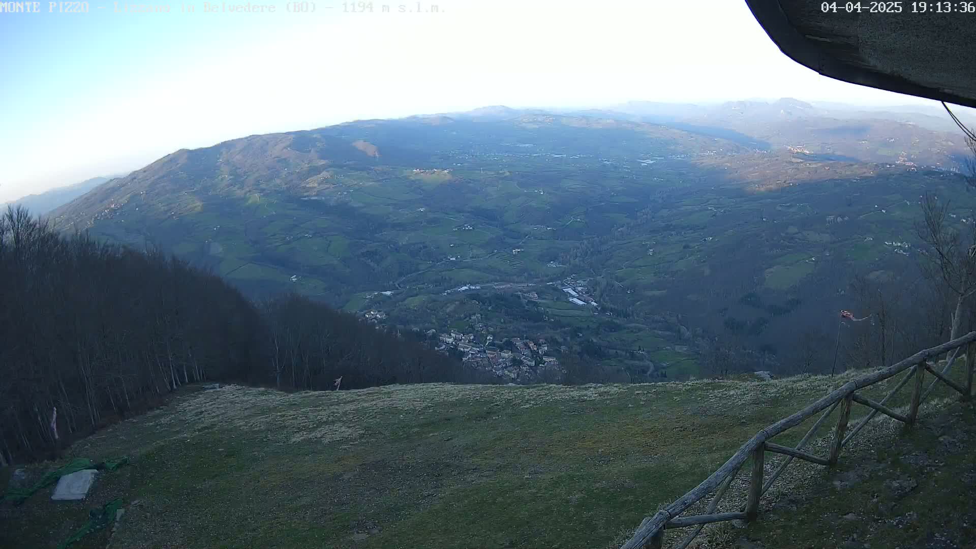 A high-angle, long shot shows a vast, hilly landscape under a clear, light-blue sky, with a small village nestled in the valley below and a wooden fence in the foreground.