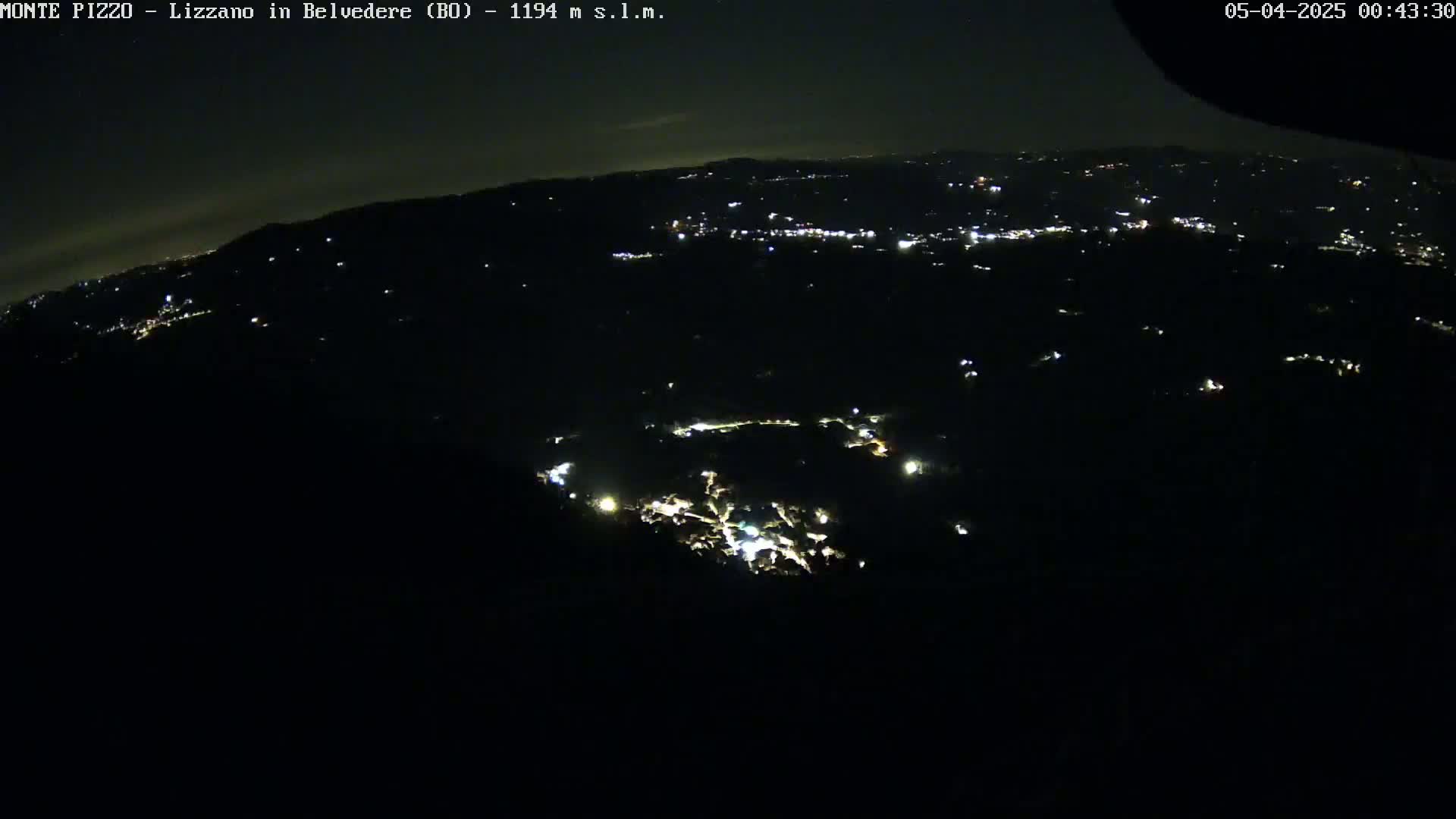 A nighttime, high-angle view shows a dark, hilly landscape sparsely dotted with small, distant lights.