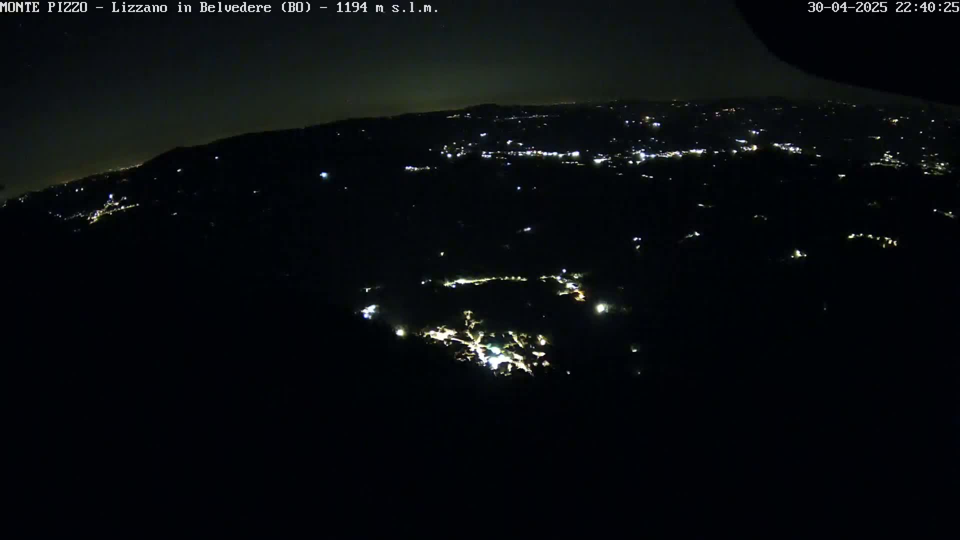 A nighttime, high-angle view shows a dark landscape with scattered lights from a town below.
