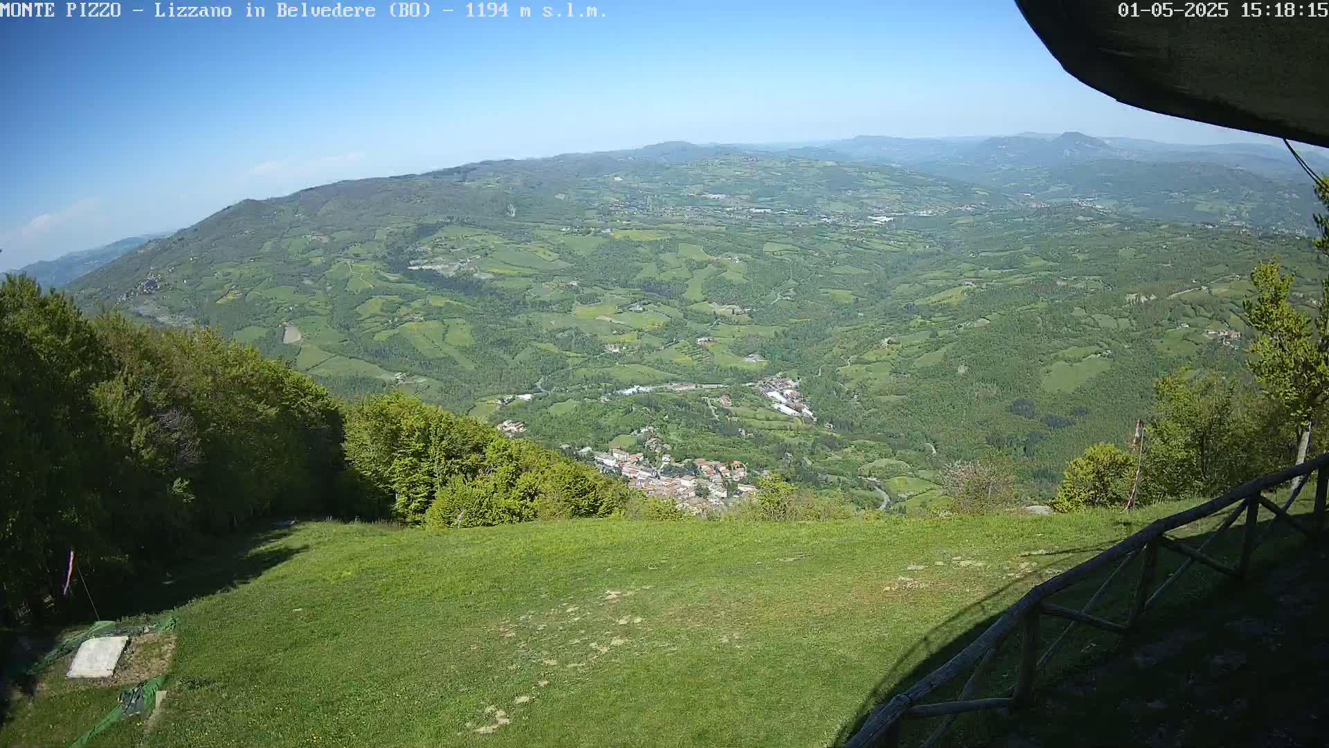 A panoramic view from a grassy hilltop shows a sprawling valley of green fields and trees under a clear blue sky.