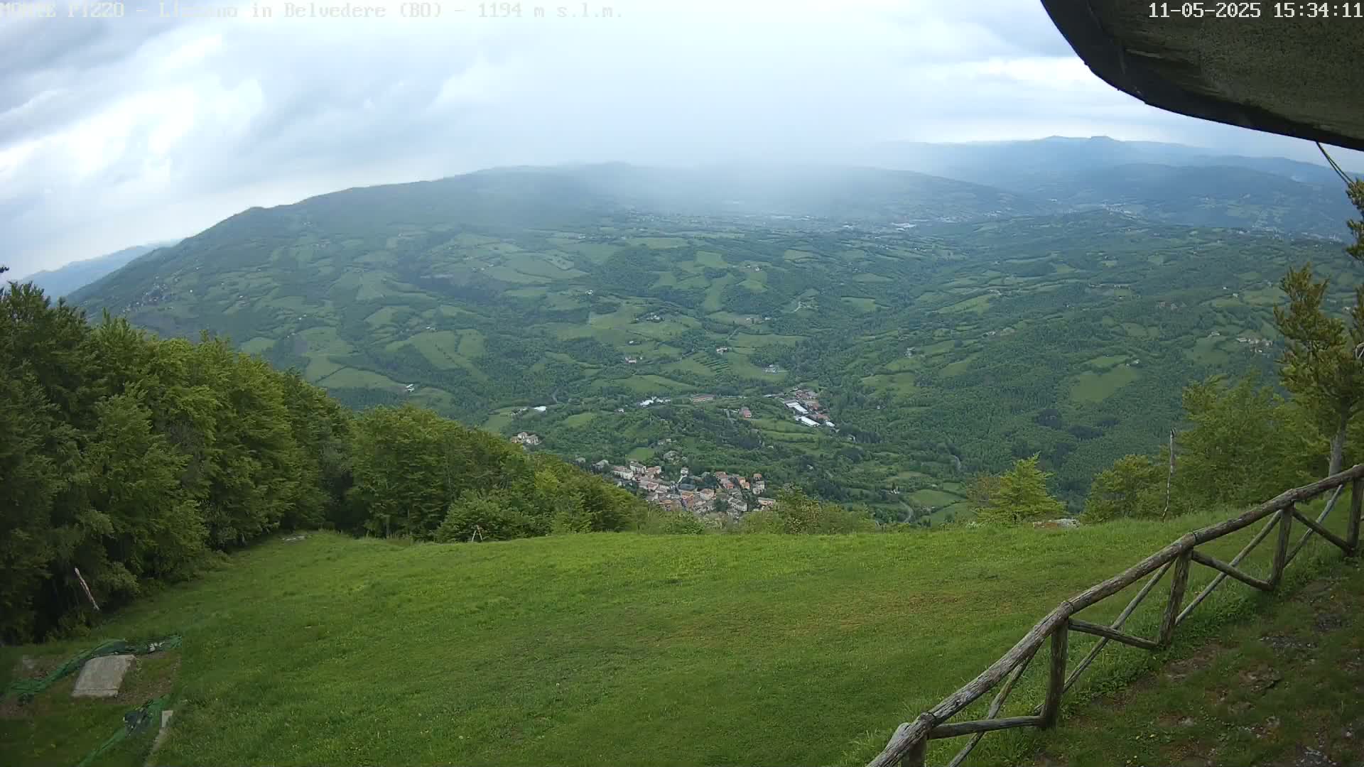 A sprawling green valley dotted with a small town is visible from a high vantage point on an overcast day, with a grassy area and wooden fence in the foreground.