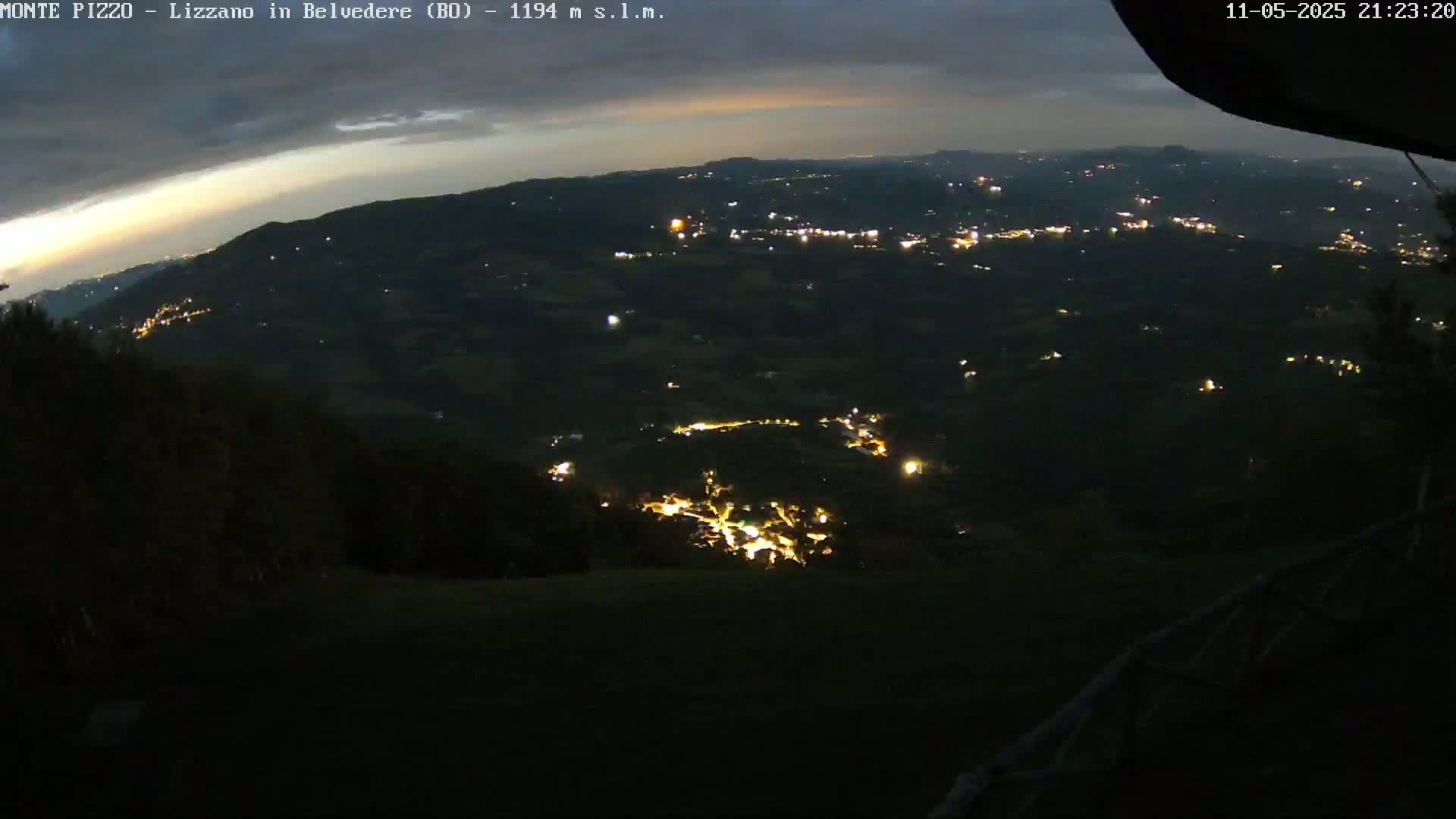 A nighttime view from a hilltop shows a valley below speckled with the lights of a town and surrounding buildings, under a partly cloudy sky.