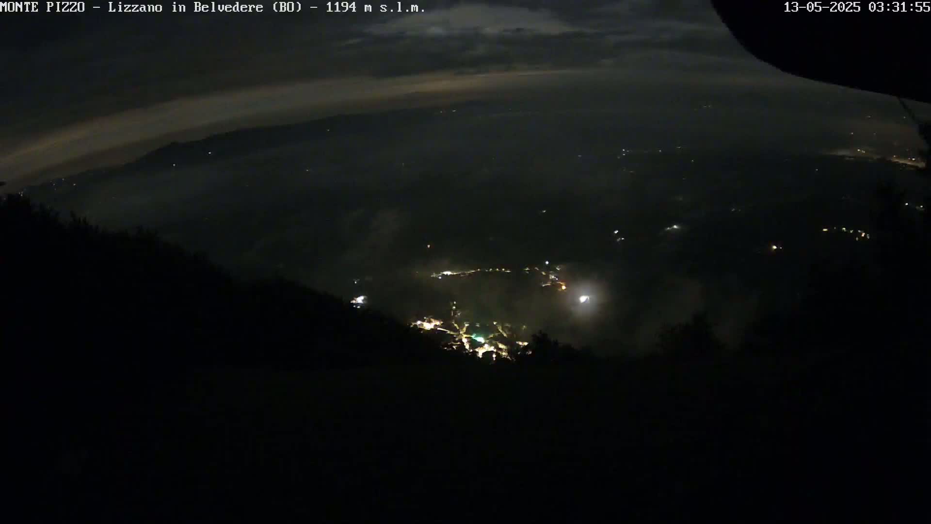 A nighttime, high-angle view shows a valley filled with faint lights, partially obscured by haze or fog under a slightly cloudy sky.