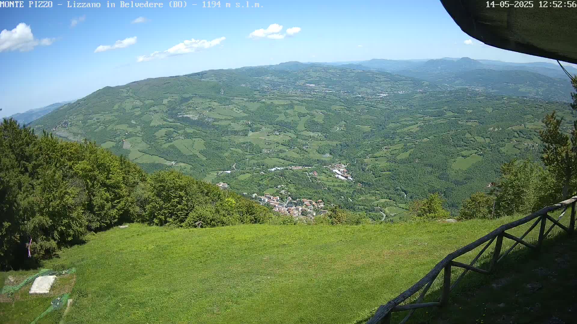 A lush green valley dotted with small towns and fields stretches out under a mostly sunny sky, viewed from a grassy hilltop with a wooden fence in the foreground.