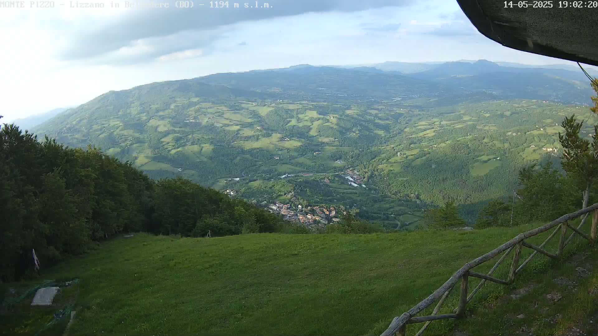 A partly cloudy daytime view from a grassy hilltop shows a vast expanse of rolling green hills and a small town nestled in the valley below, with a wooden fence in the foreground.