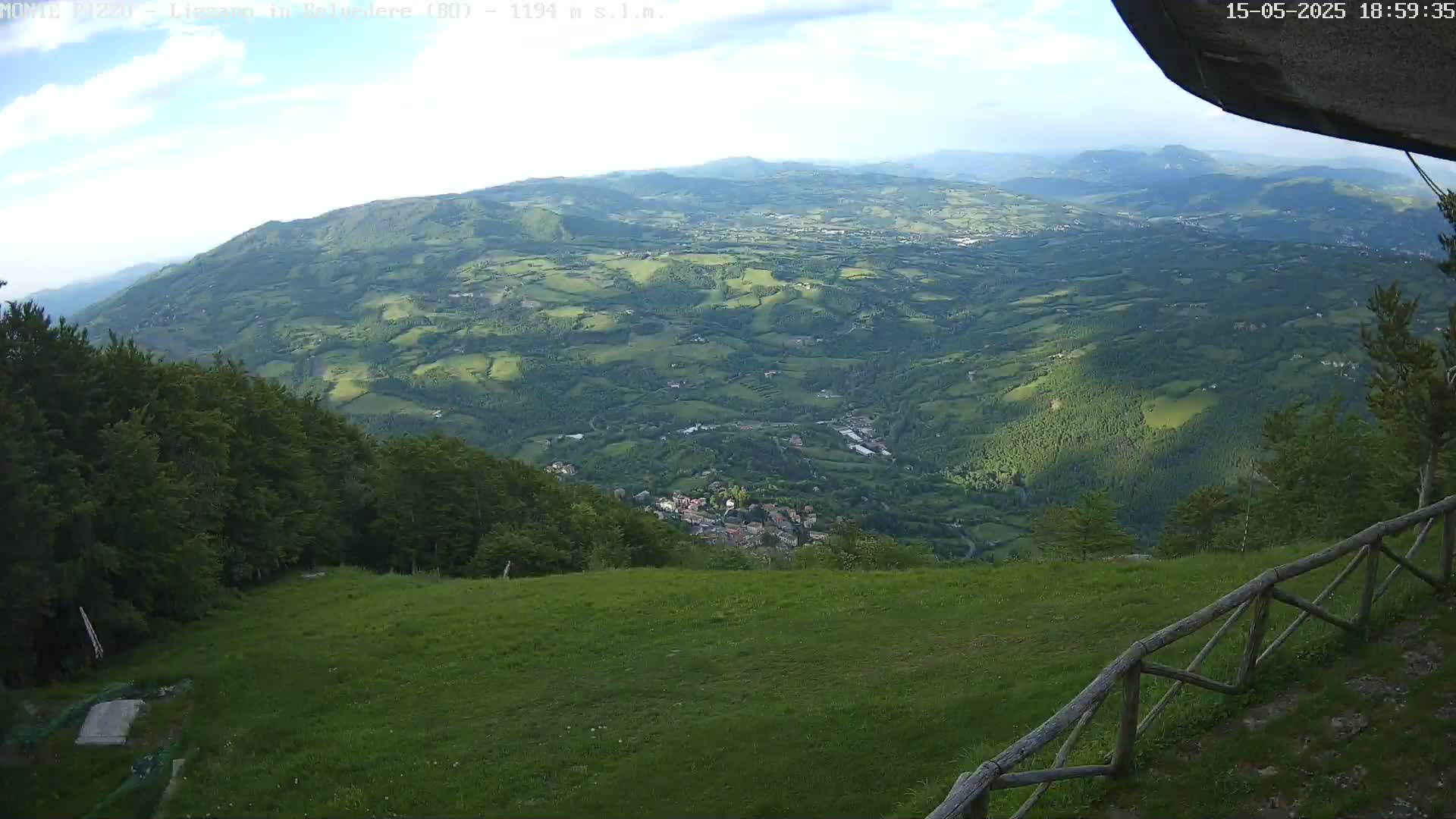 A partly cloudy daytime view from a grassy hillside shows a valley of rolling green hills and a small town below, with a wooden fence in the foreground.