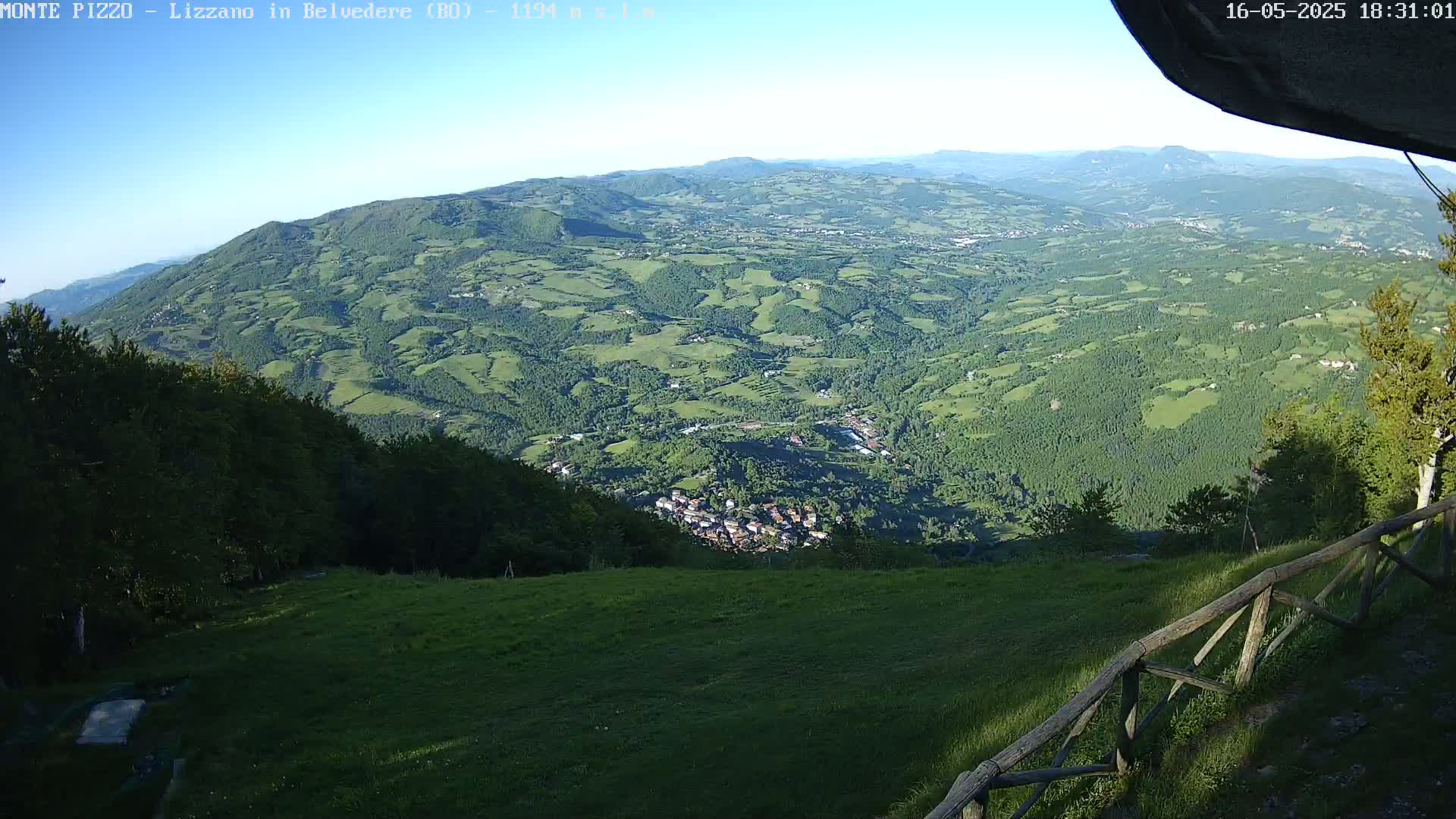 A panoramic view from a hilltop shows a vast, verdant valley speckled with small towns and farms under a clear, sunny sky.