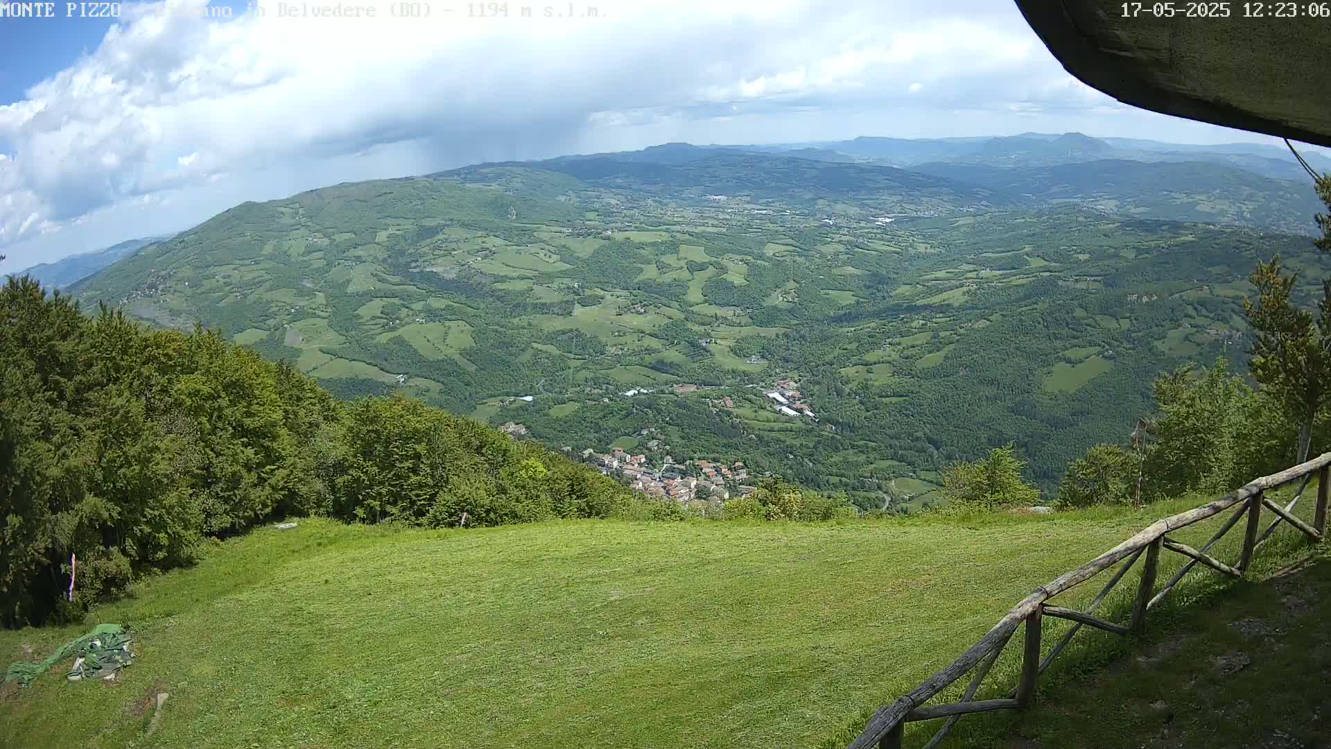 A grassy area with a wooden fence overlooks a valley of green hills and a small town under partly cloudy skies.
